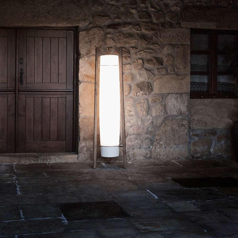 Lit column lamp with wooden supports and grey bottom cap beside a wood door and window in a rustic stone wall