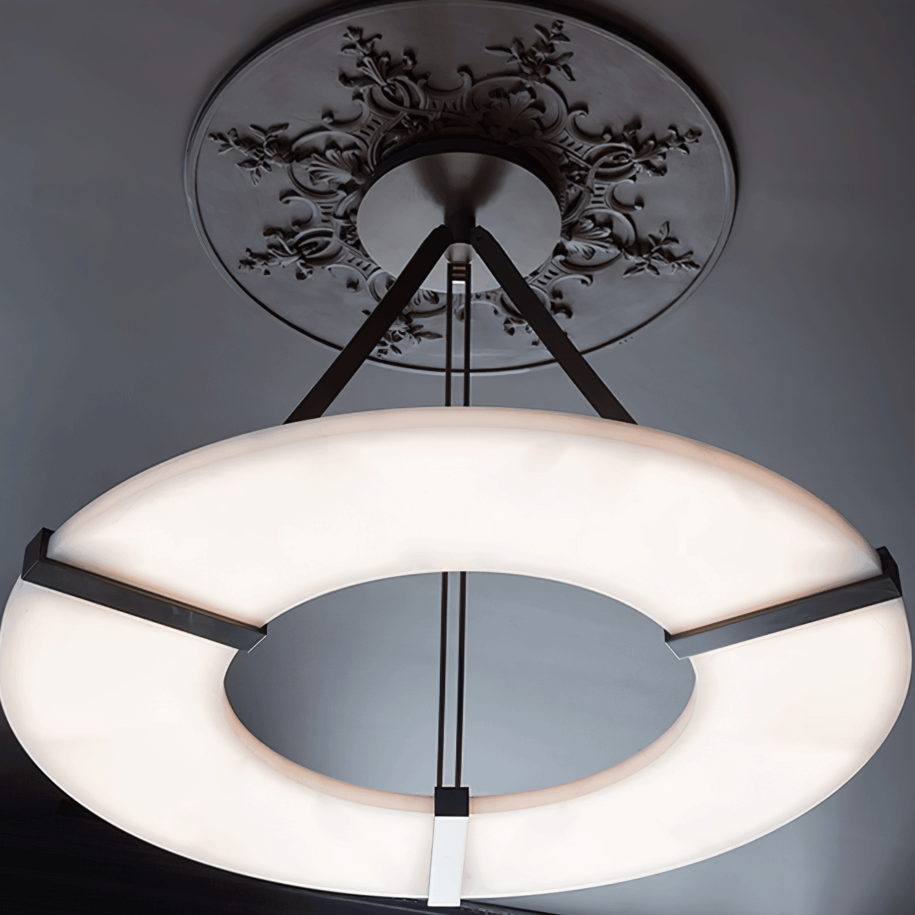 Close underside of lit white ring, black brackets and supports beneath floral ceiling ornament