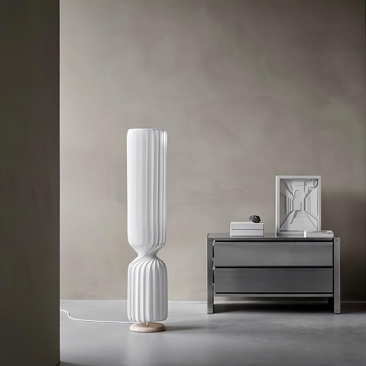 White Twist lamp beside gray chest with framed perspective drawing and books, with white floor cord and round base