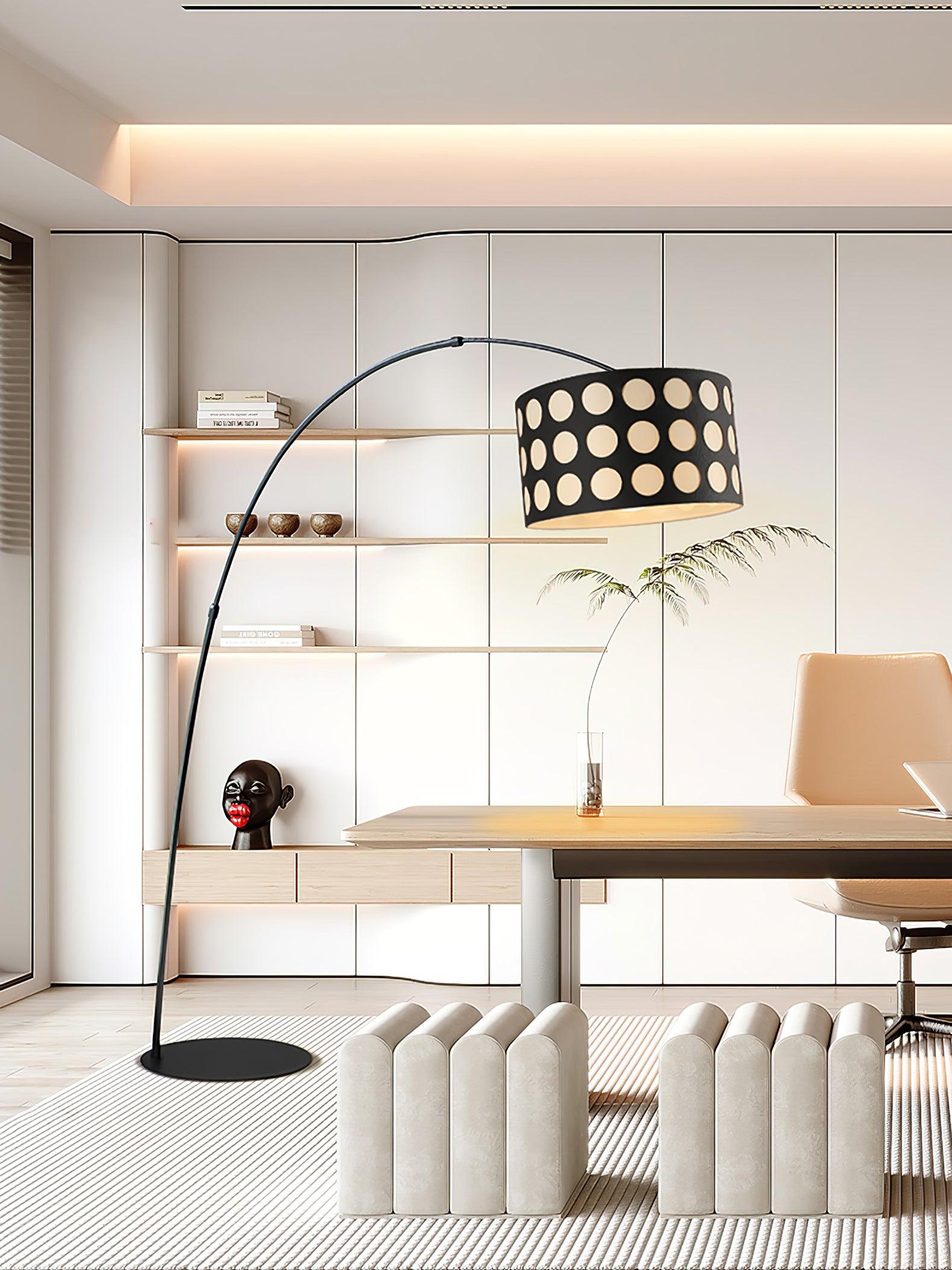 Black arc floor lamp with dotted drum shade beside desk, pale chair and sculptural upholstered bench