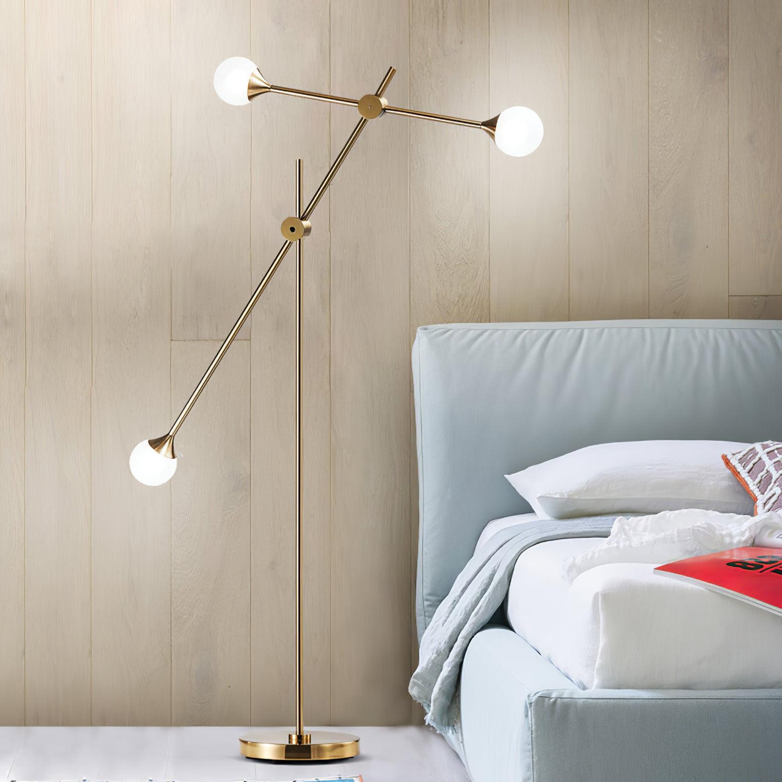 Three-globe gold Kemi floor lamp beside a pale blue upholstered bed against a wood-panelled wall