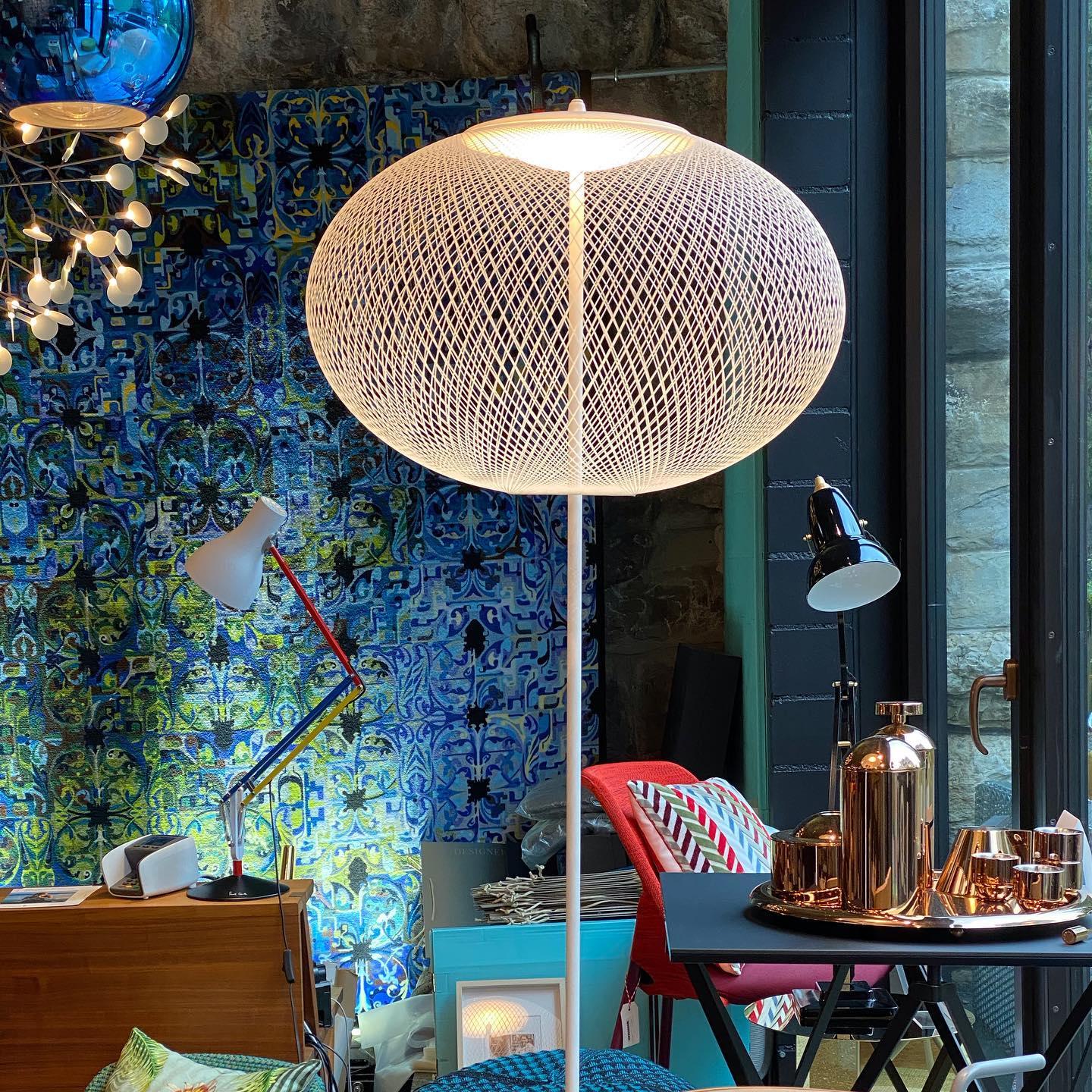 Close view of a glowing white mesh floor lamp in a colorful showroom with blue patterned fabric and copper-tone tableware