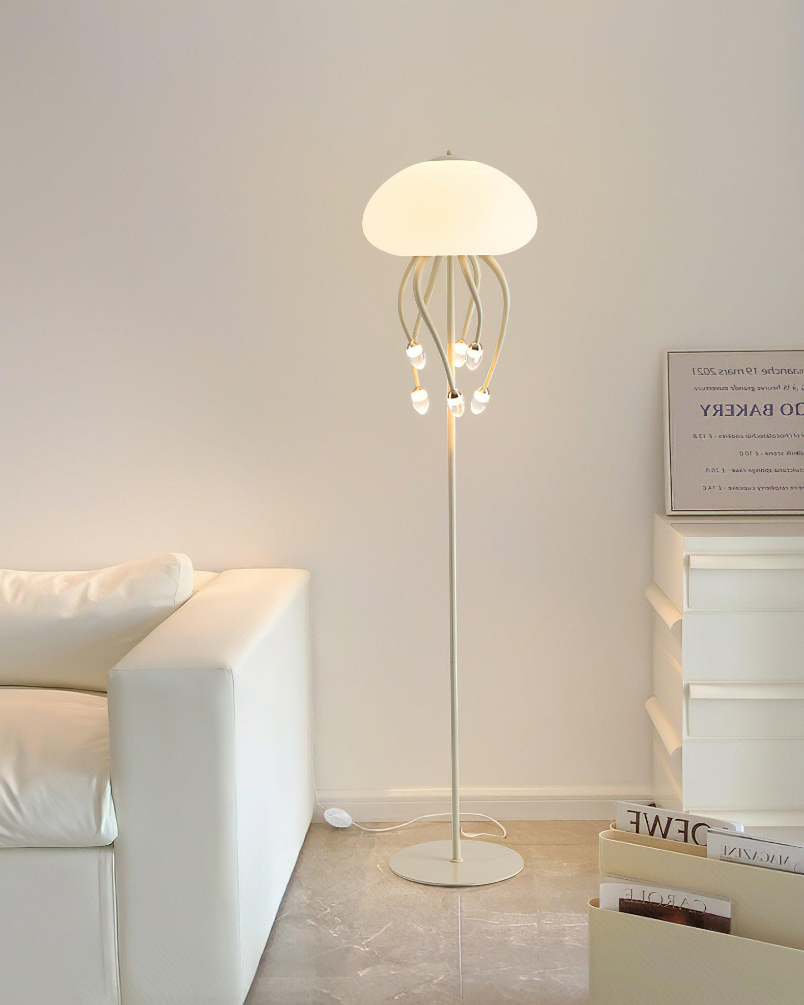 Beige six-arm floor lamp in sofa-and-drawers corner, with glowing dome, hanging droplets, slender pole and round base.