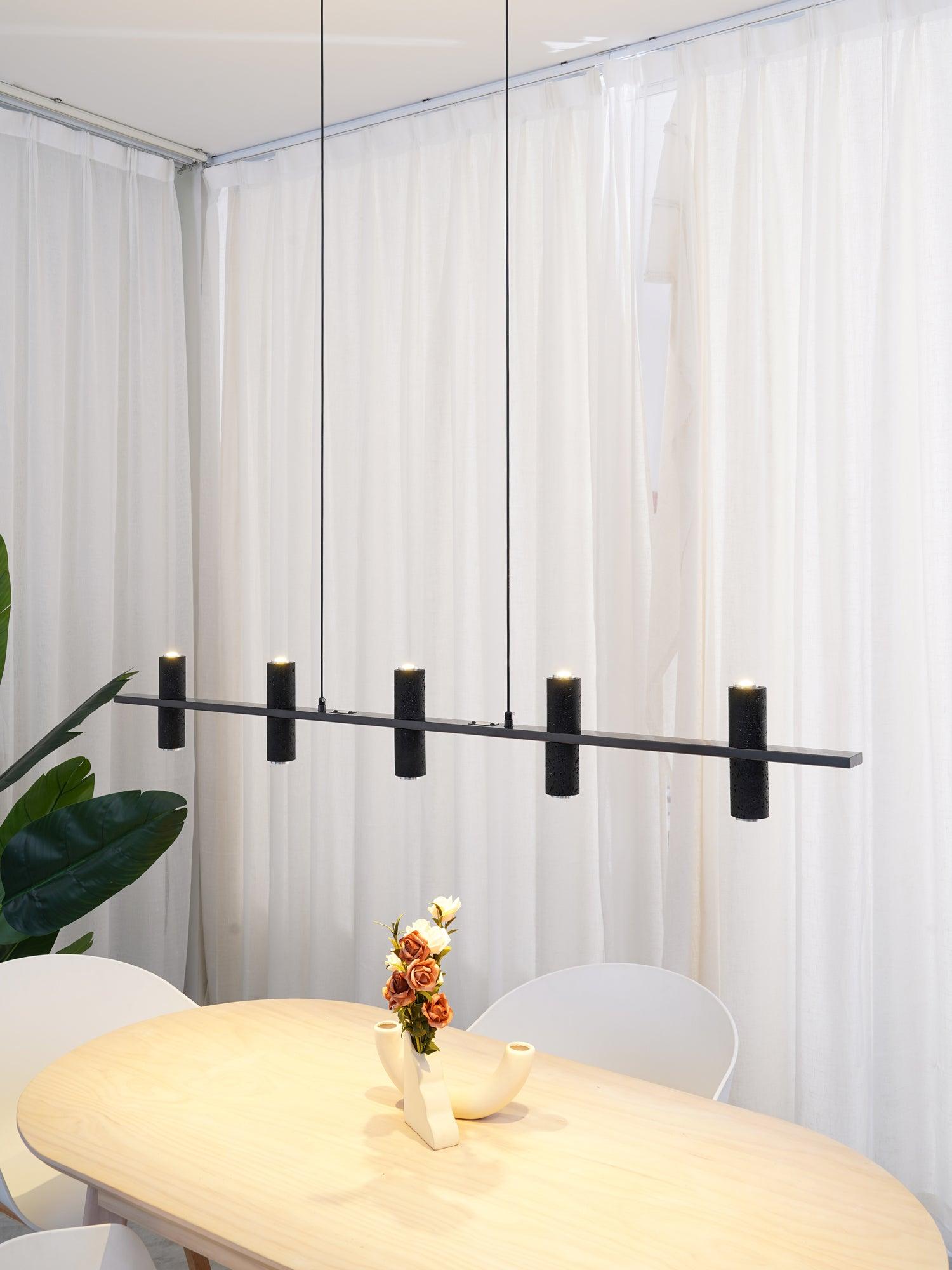Five black stone cylinders with lit upper openings over pale table, cream branching vase, orange flowers and green leaves.