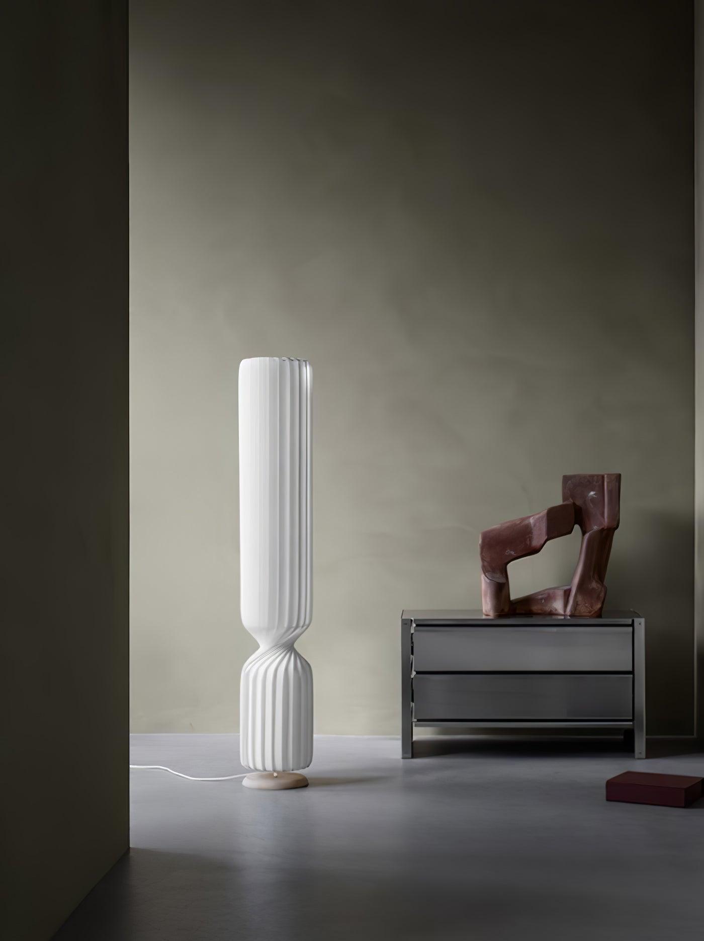 Unlit-looking white Twist lamp by gray metal chest and reddish sculpture, with red object on floor and cord extending left