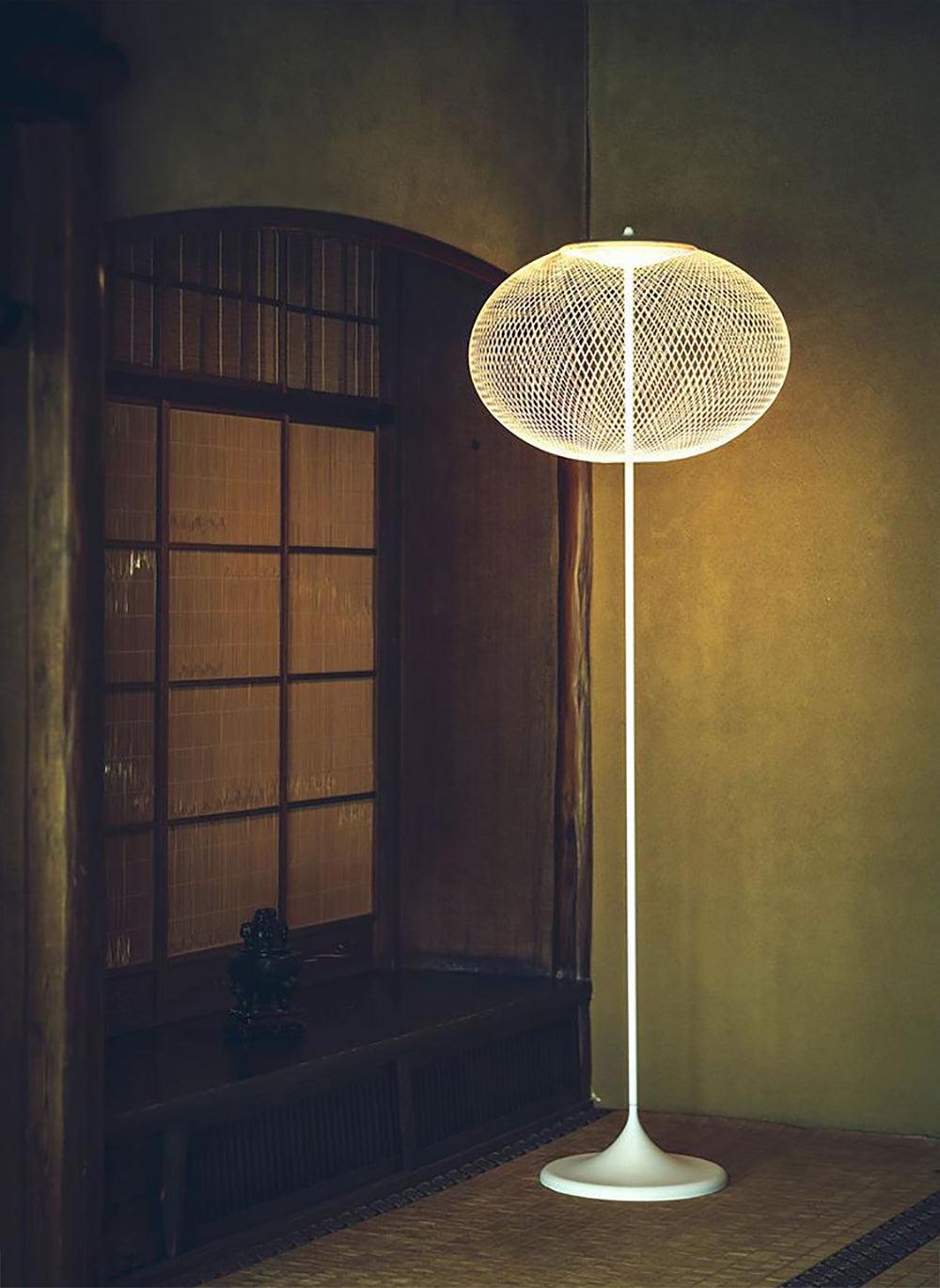 White mesh floor lamp glowing beside wood-framed panels and a woven floor mat in a dim interior