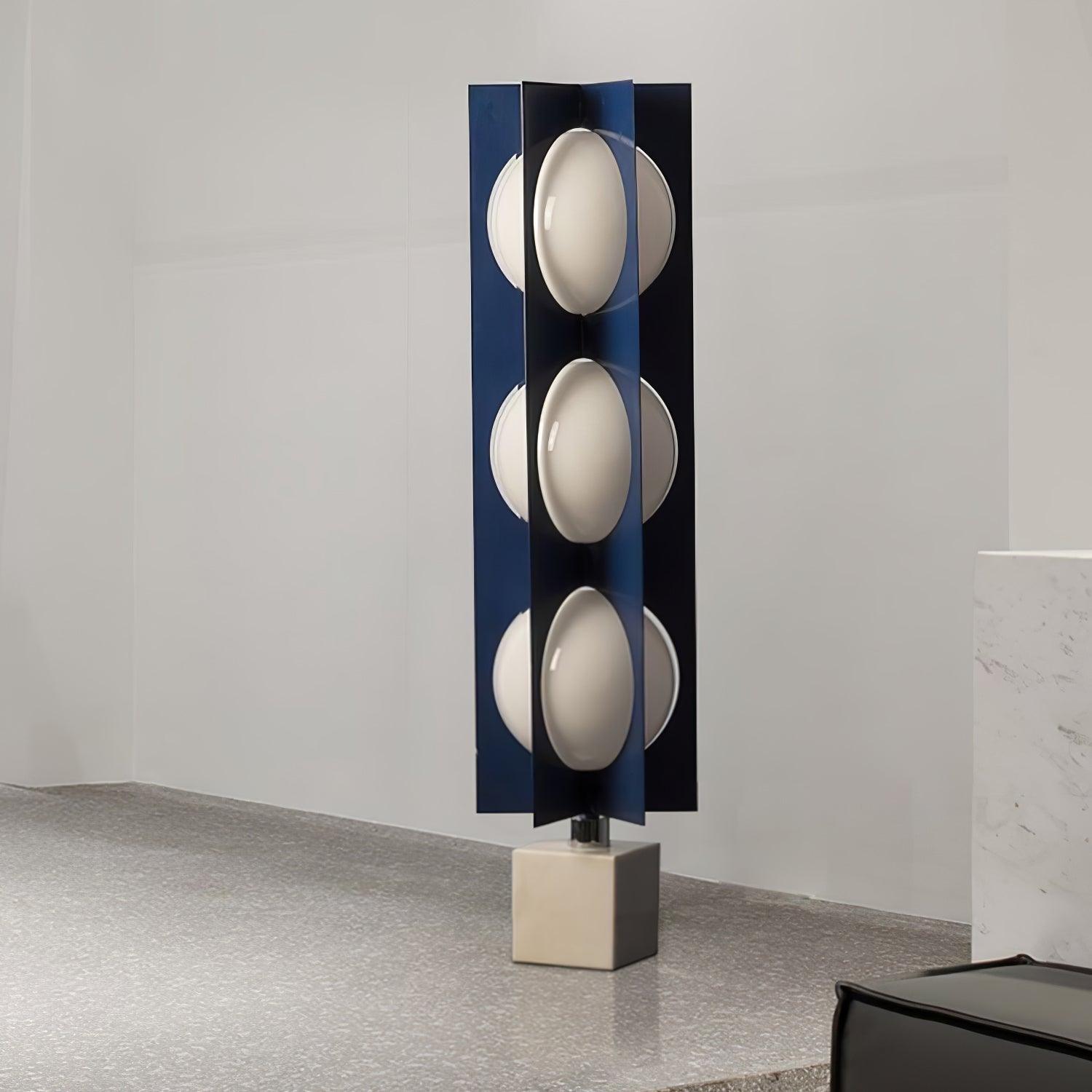 Unlit blue column lamp with three white shade levels and a marble block on a stone floor beside a pale plinth
