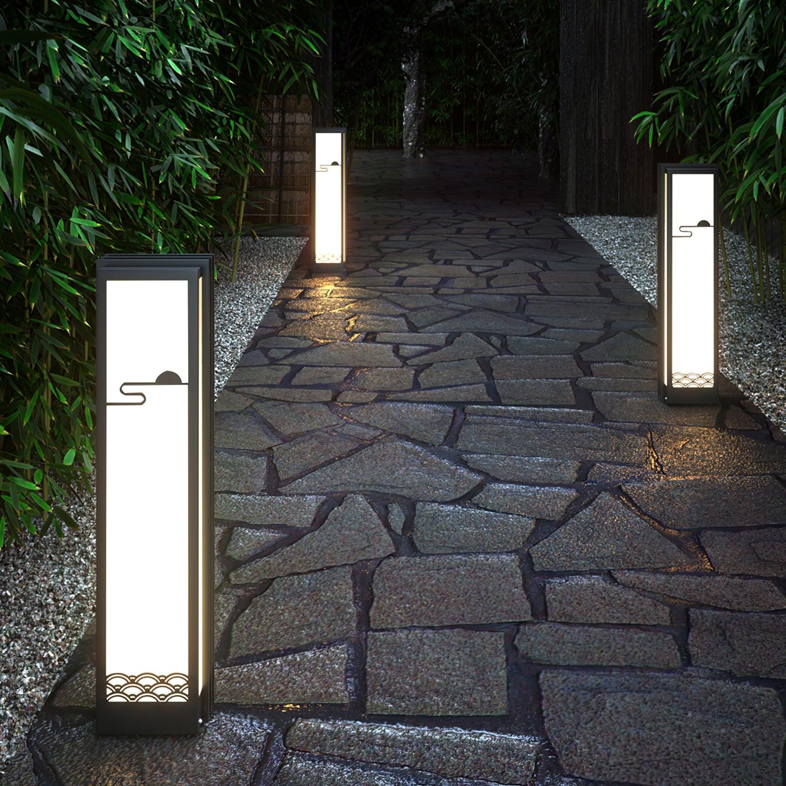 Tall slim garden lanterns along a rough stone path between bamboo, trees and gravel borders at night