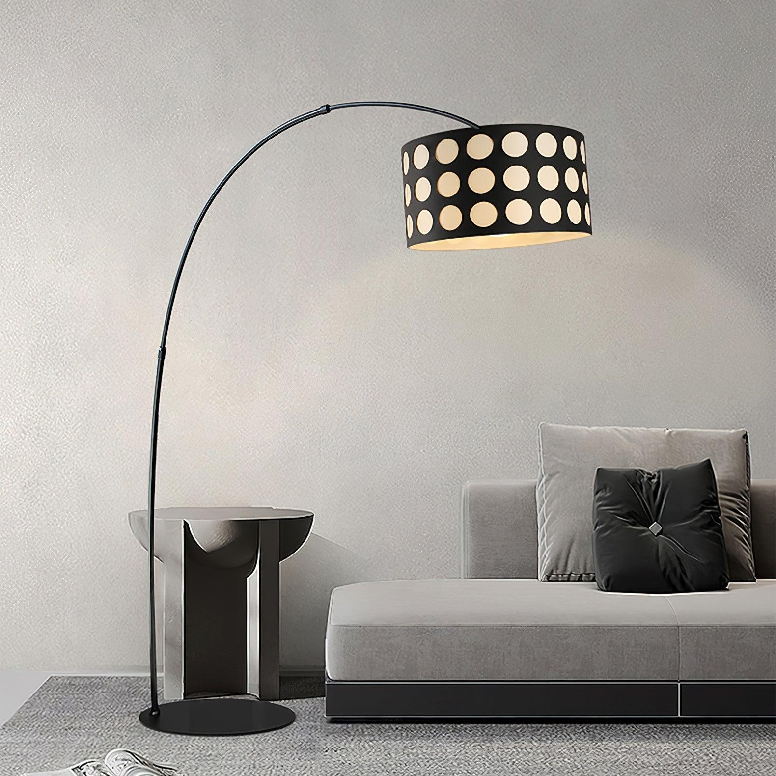 Black arc floor lamp with dotted shade above pale sofa beside reflective side table, black cushion and textured wall