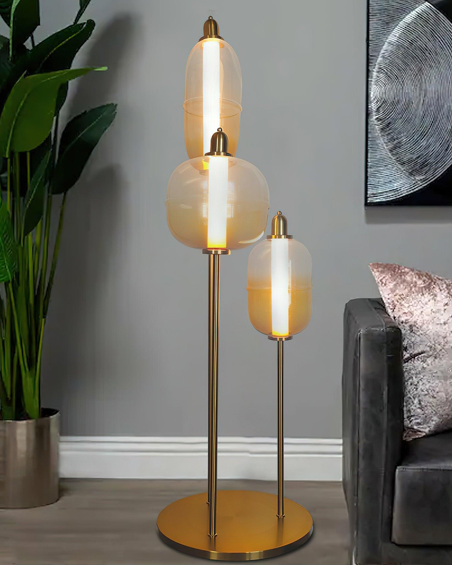 Closer view of the staggered amber shades and gold round base beside a plant, dark sofa and pink cushion