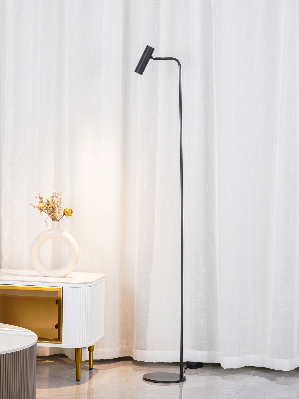 Portrait black floor lamp with tilted cylindrical head beside white curtains, ring vase and white cabinet with gold inset