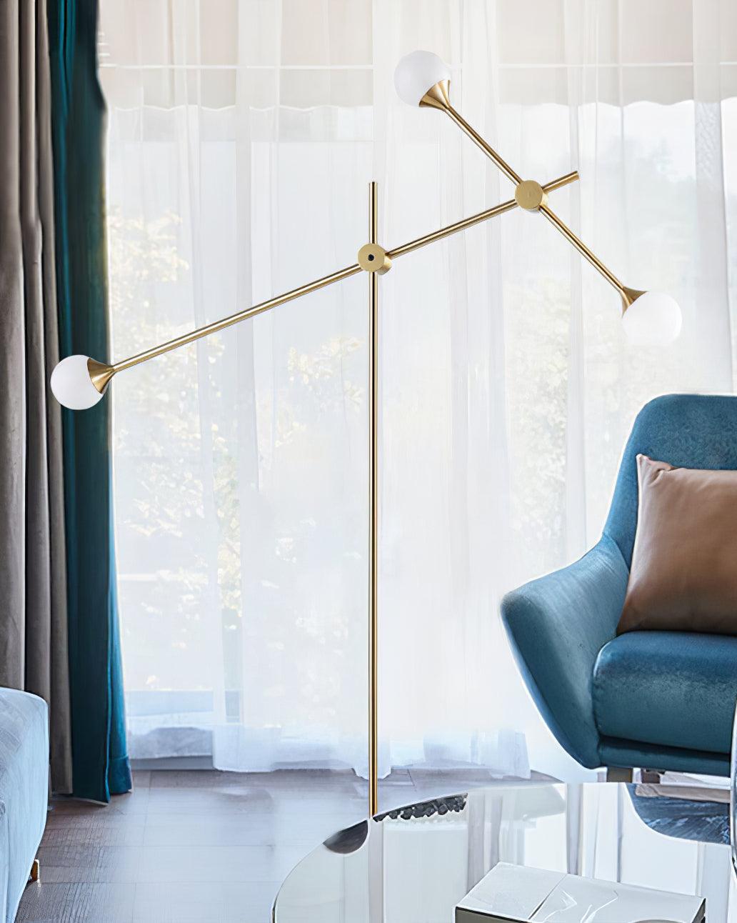 Kemi lamp with crossing gold arms and three white globes by sheer curtains, a blue armchair and glass coffee table