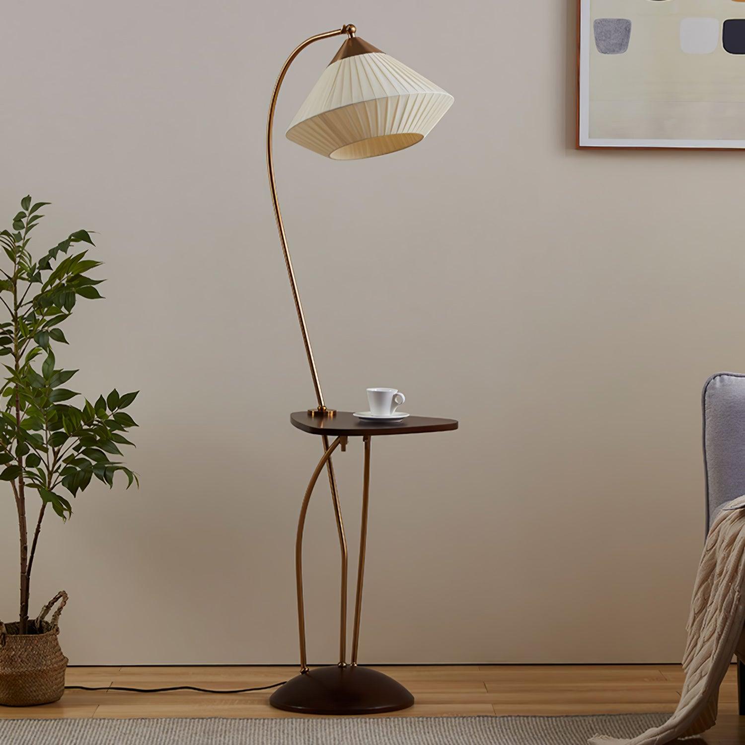 Unlit Curve lamp showing its beige pleated shade, curved supports, cup-topped dark table and domed base by a grey chair