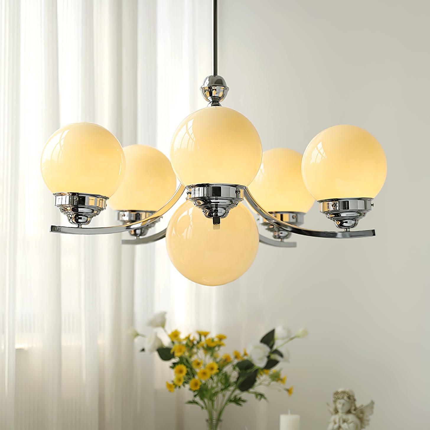 Warm-looking glass globes on rising chrome arms above flowers and candles beside white curtains