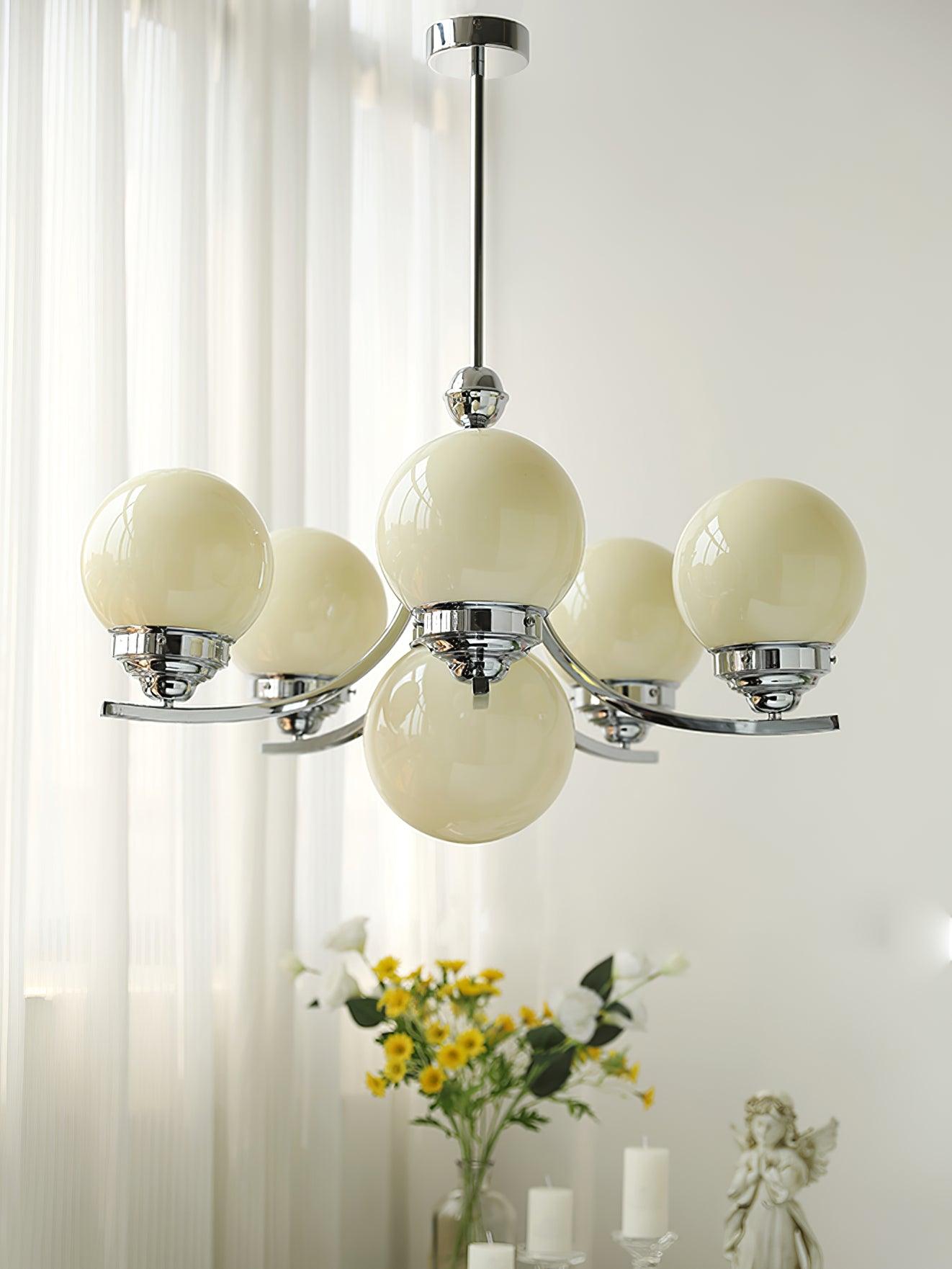 Larger rising-arm Dewdrop with pale beige-looking globes beside white curtains, flowers and a figurine
