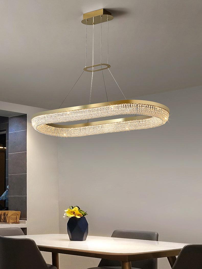 Long rounded-oval gold crystal chandelier over a rectangular dining table with blue vase and yellow flowers