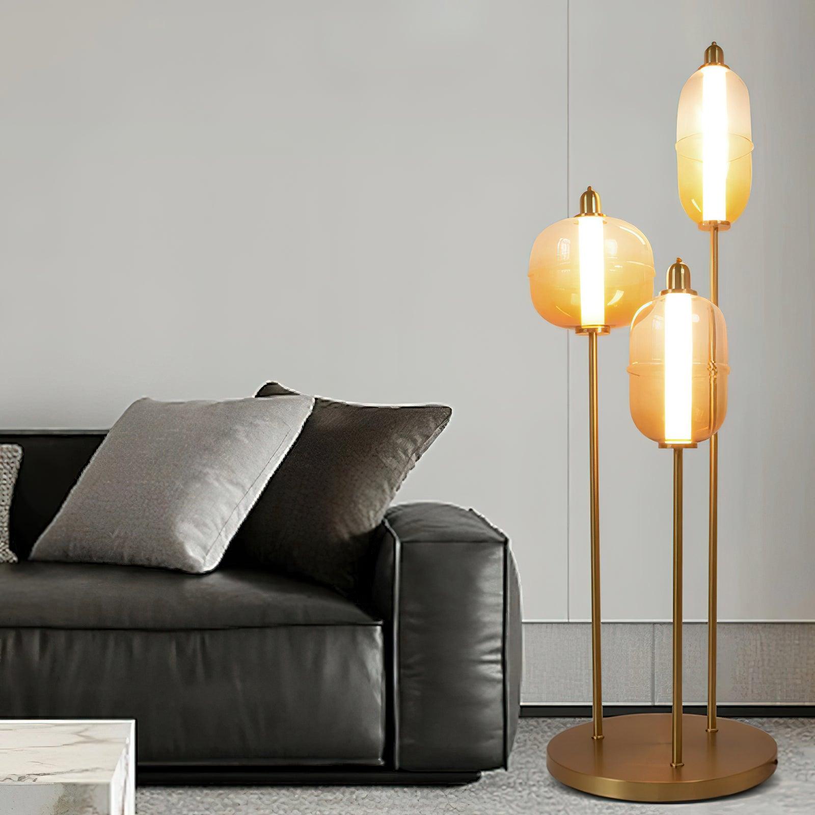 Amber glass floor lamp with three staggered shades and round gold base beside a dark sofa with gray cushions