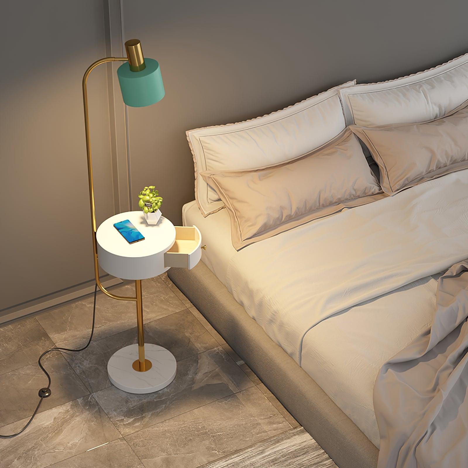 High view of gold lamp beside cream bed, green shade, white open drawer table with phone and plant, round base and floor switch