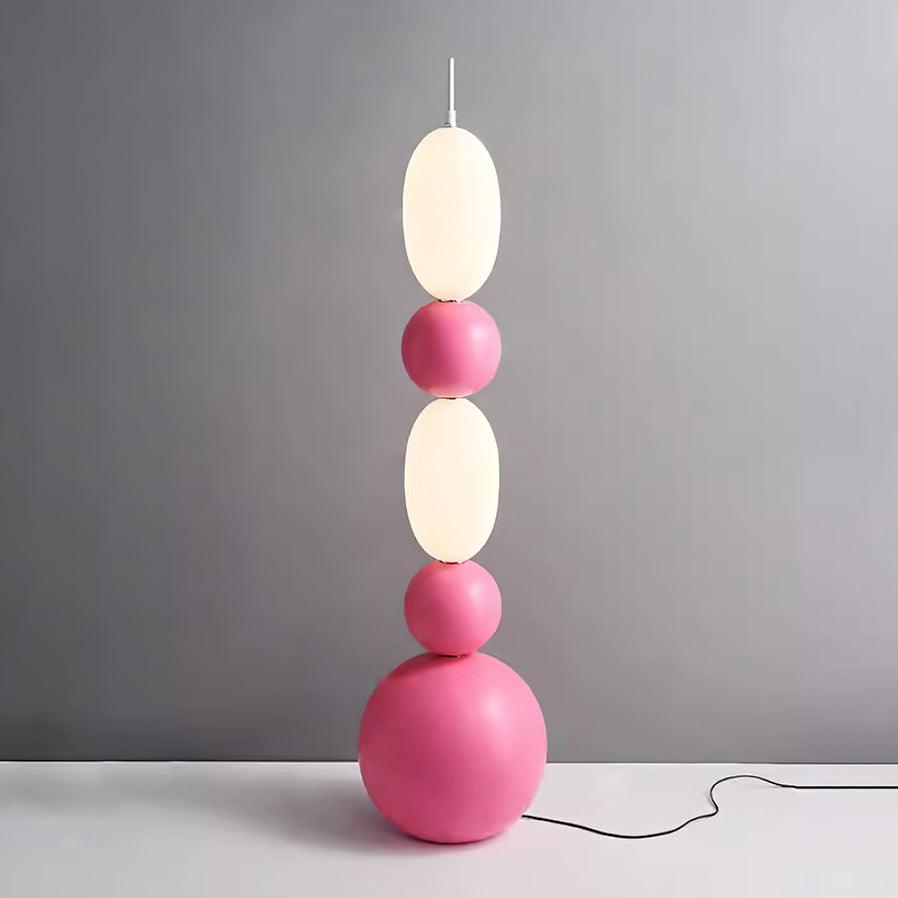 Wider view of pink Bola floor lamp with glowing white oval shades, spherical details and black cord by a grey wall