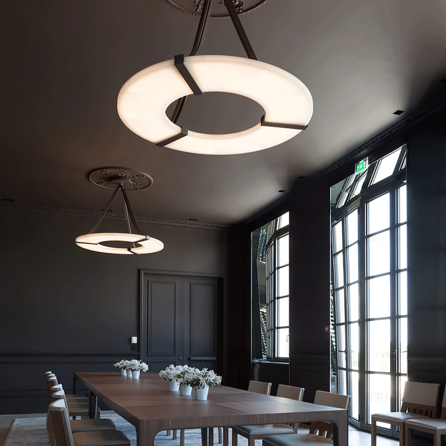 Two lit halo pendants above long wooden dining table with chairs and flower pots beside tall windows