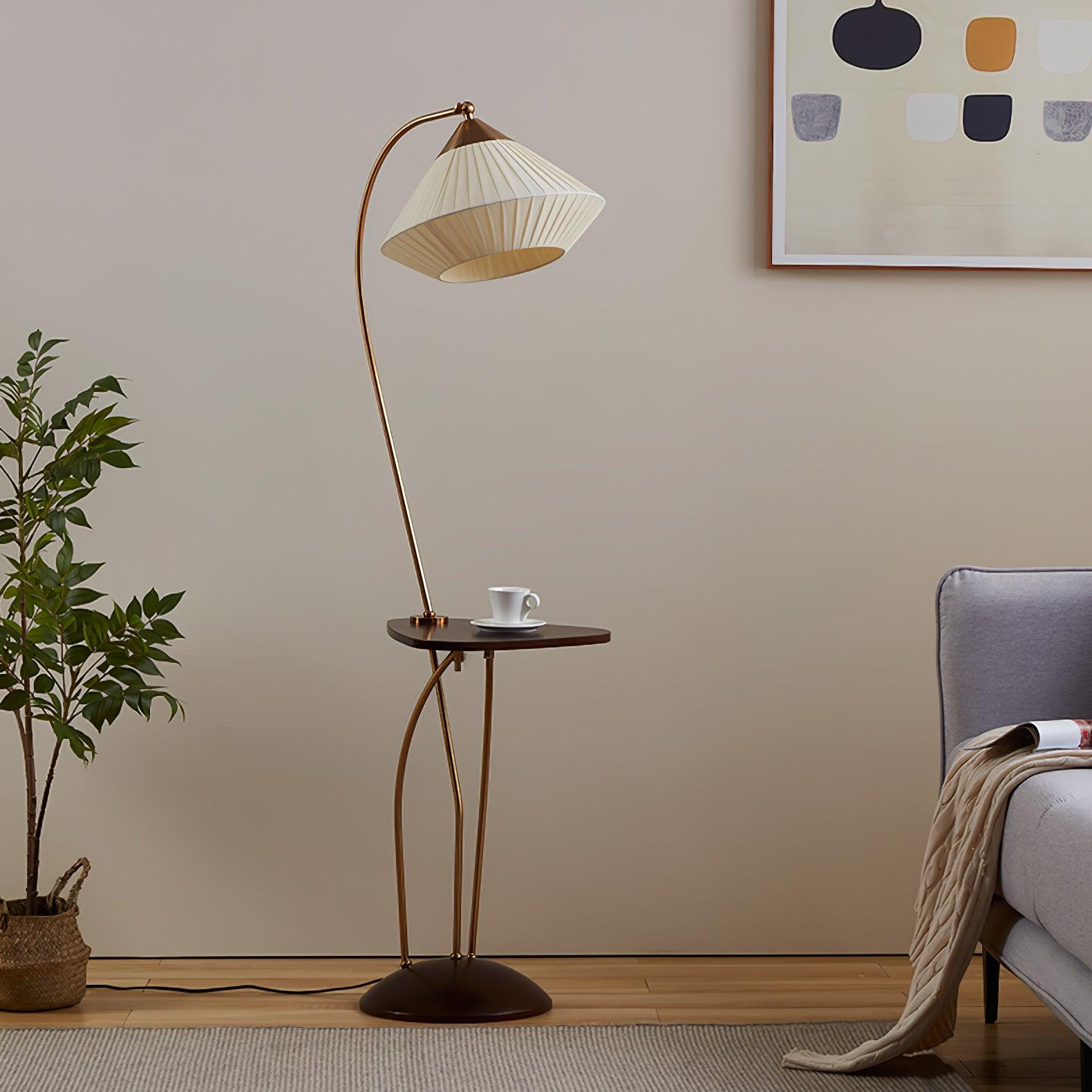 Full view of unlit Curve’s curved frame, beige pleated shade, small table and floor cable beside a grey armchair