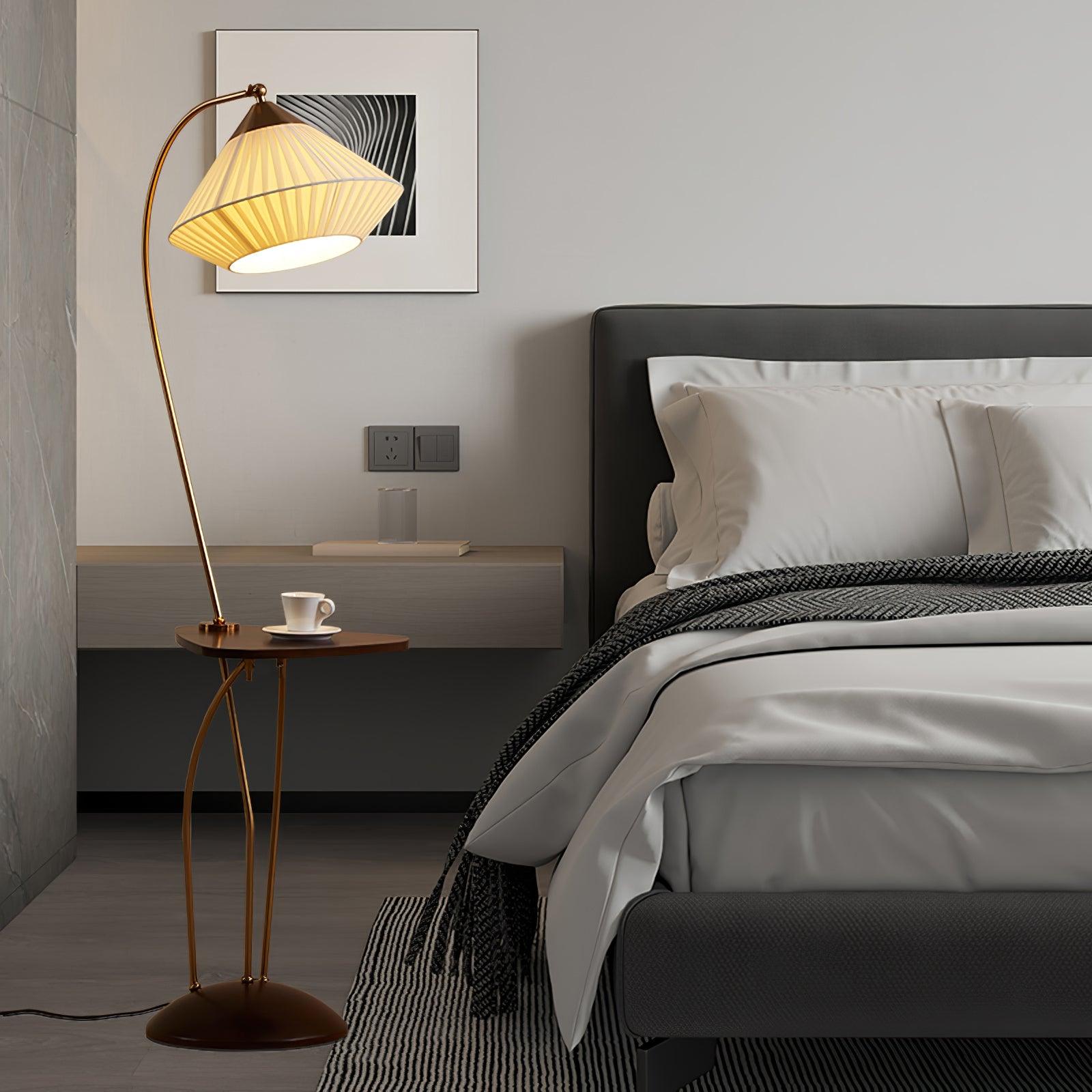 Lit Curve lamp with a small table and cup beside a bed with white pillows, dark headboard and patterned throw