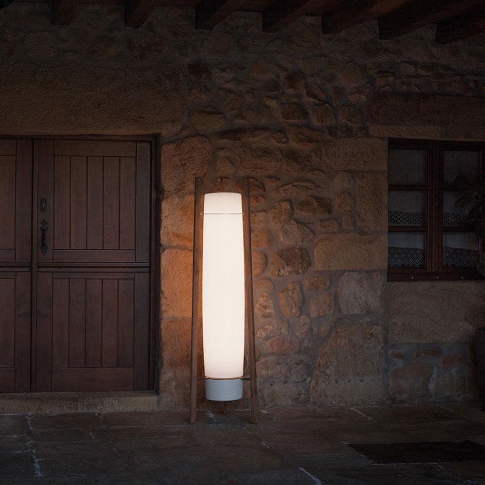 Darker view of a lit white column lamp with wooden supports and grey bottom cap against a rustic stone wall