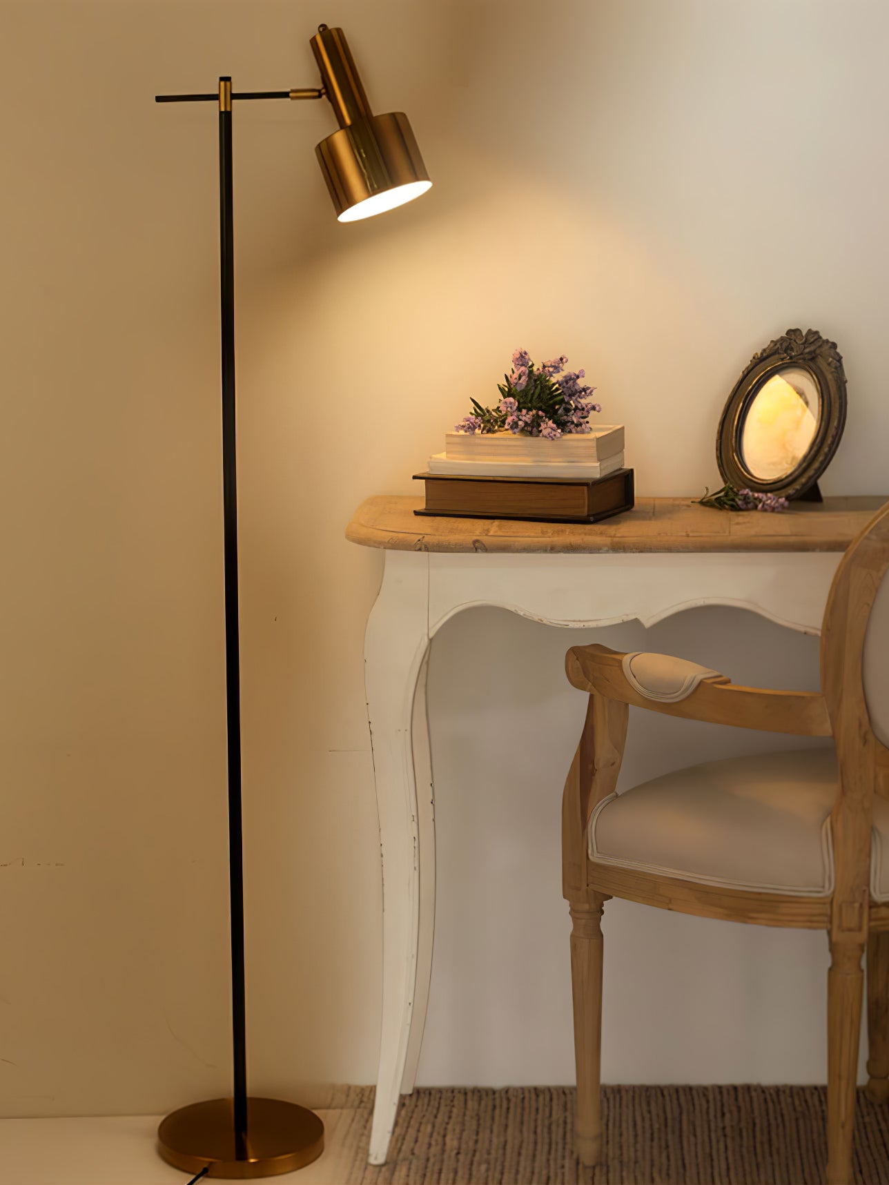 Lit gold-headed floor lamp beside white writing desk with books, flowers, framed mirror and upholstered chair