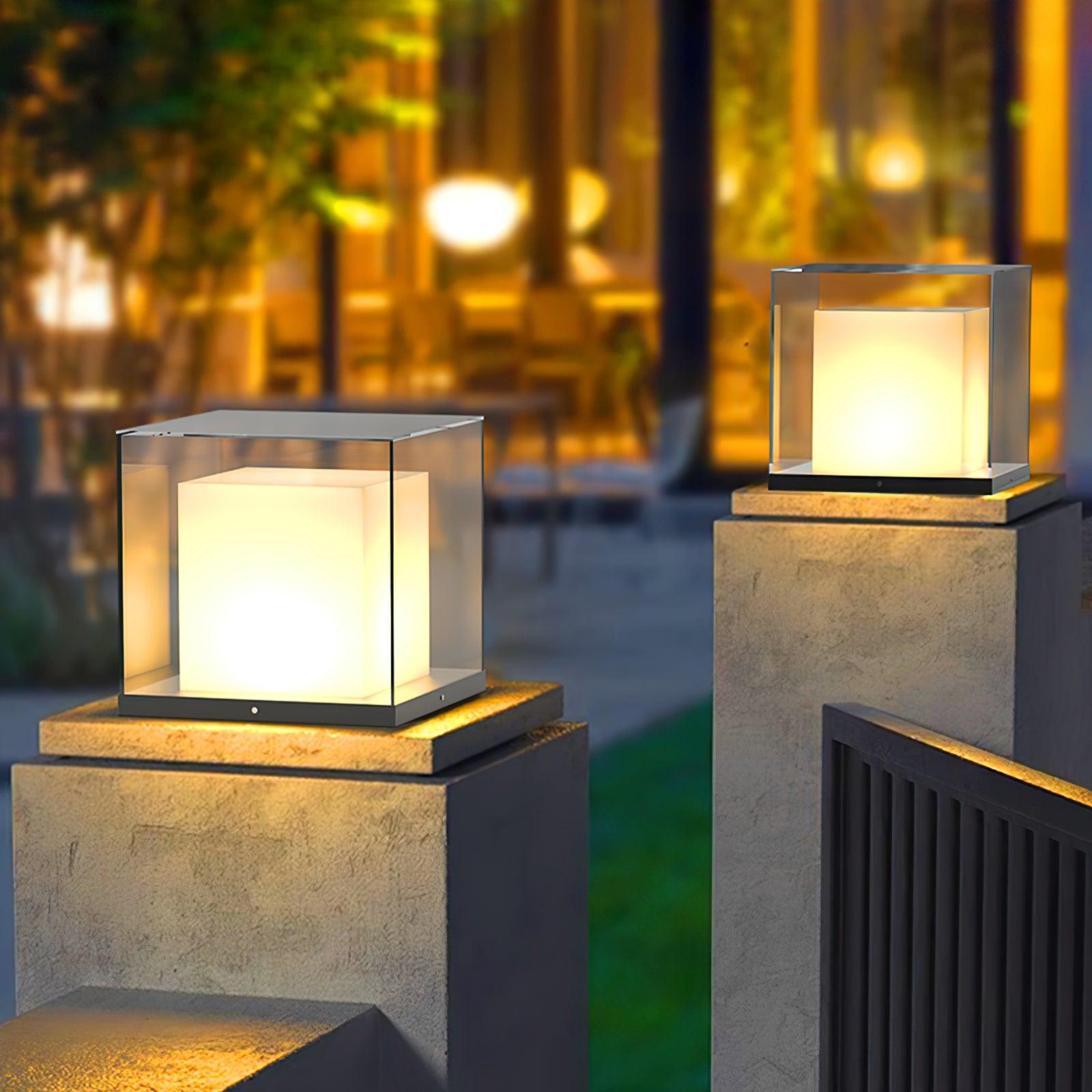 Two illuminated glass cube lights on stone-textured gate pillars beside a dark slatted gate and trees at dusk