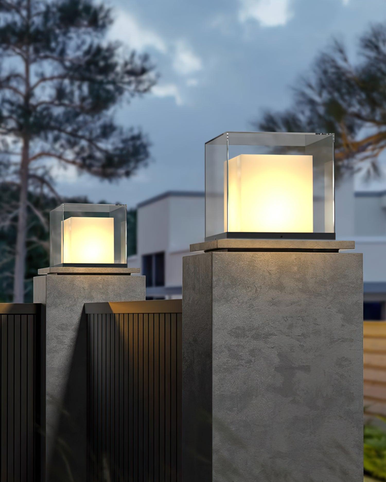 Two glowing cube lights on rough gate pillars beside a dark vertical-slat gate, white house and trees at dusk