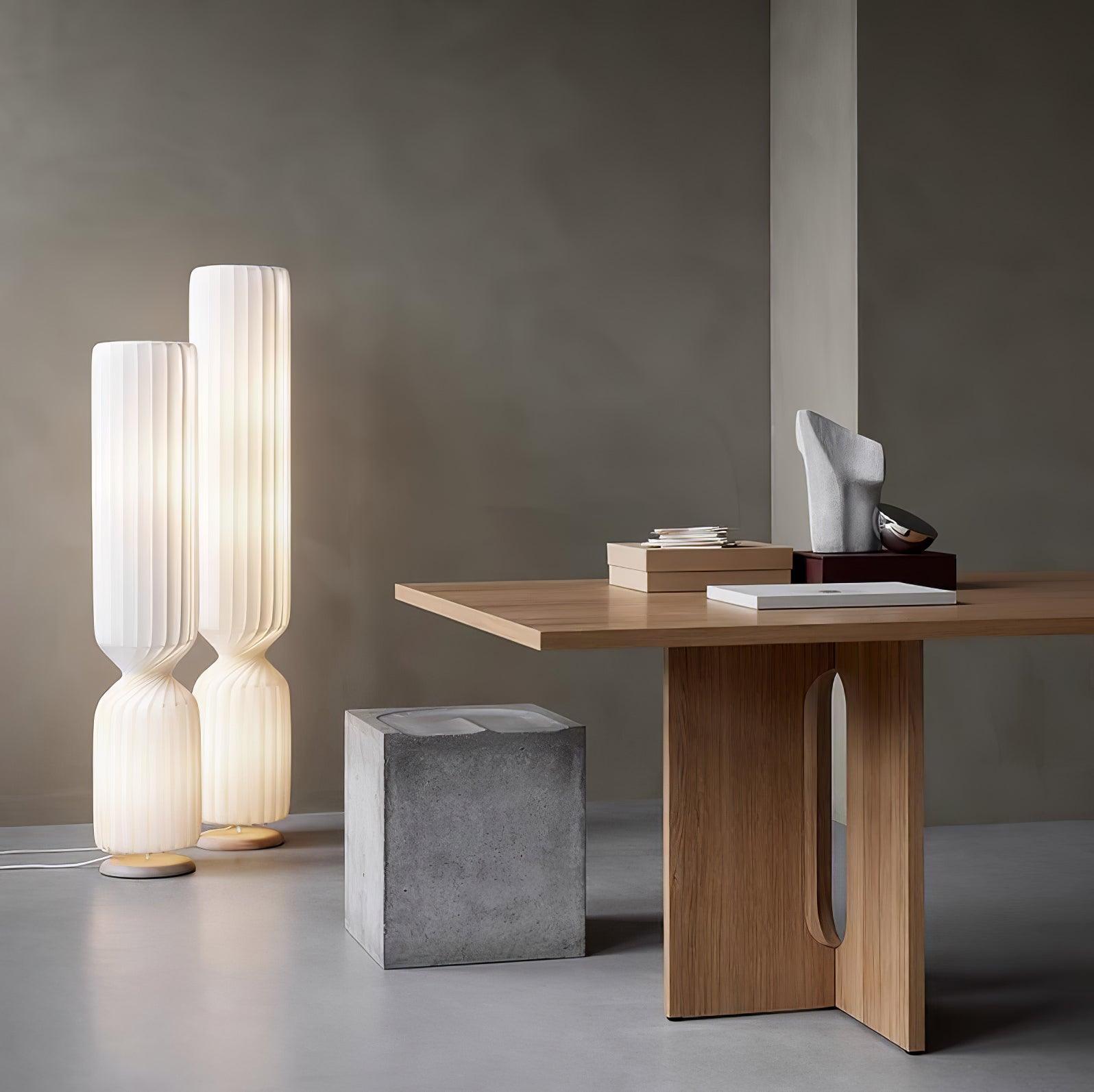 Two lit white pleated twist lamps of different heights beside wooden console, books, sculpture and gray block stool