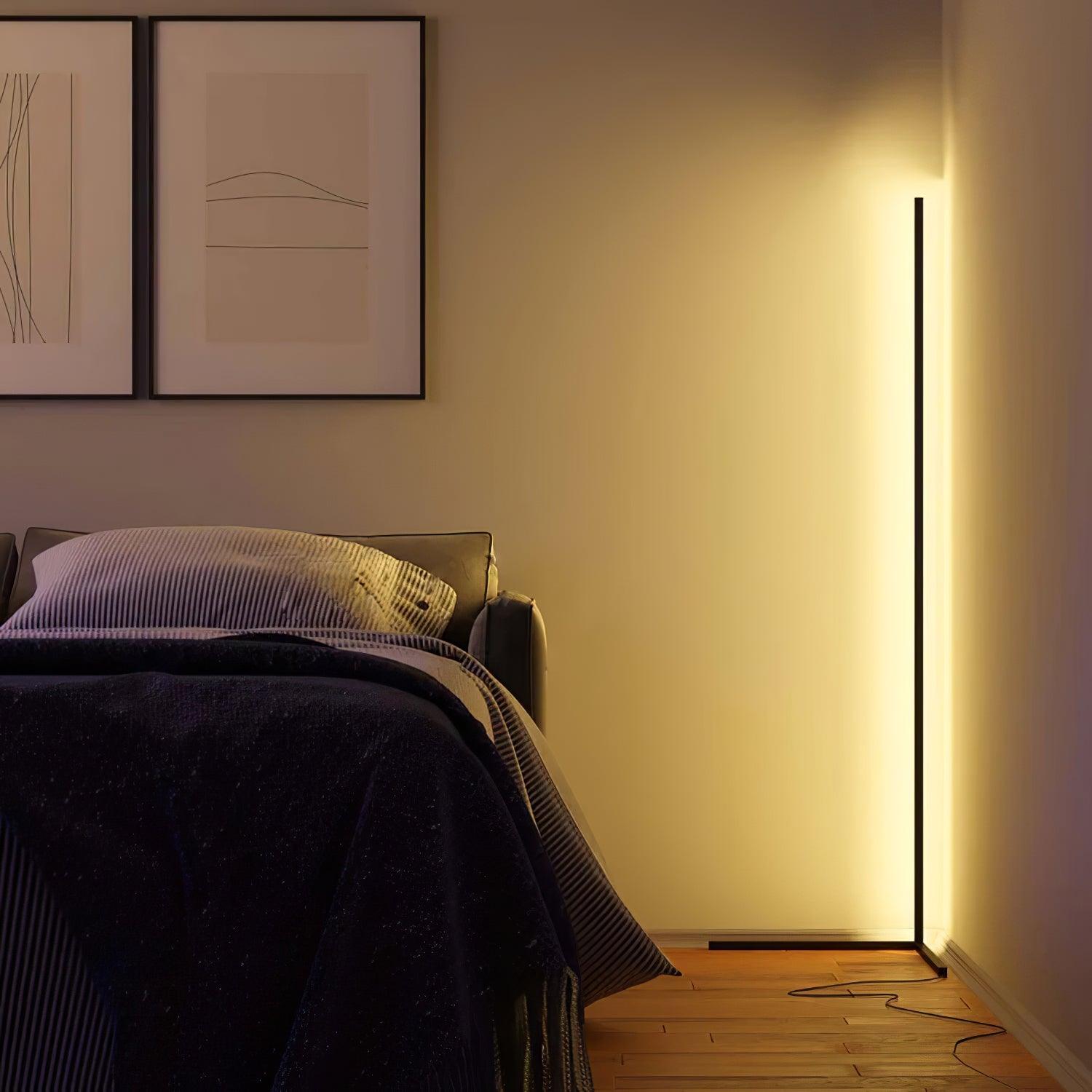 Warm-lit black floor lamp beside a bed in a dim bedroom, with a loop of power cord visible