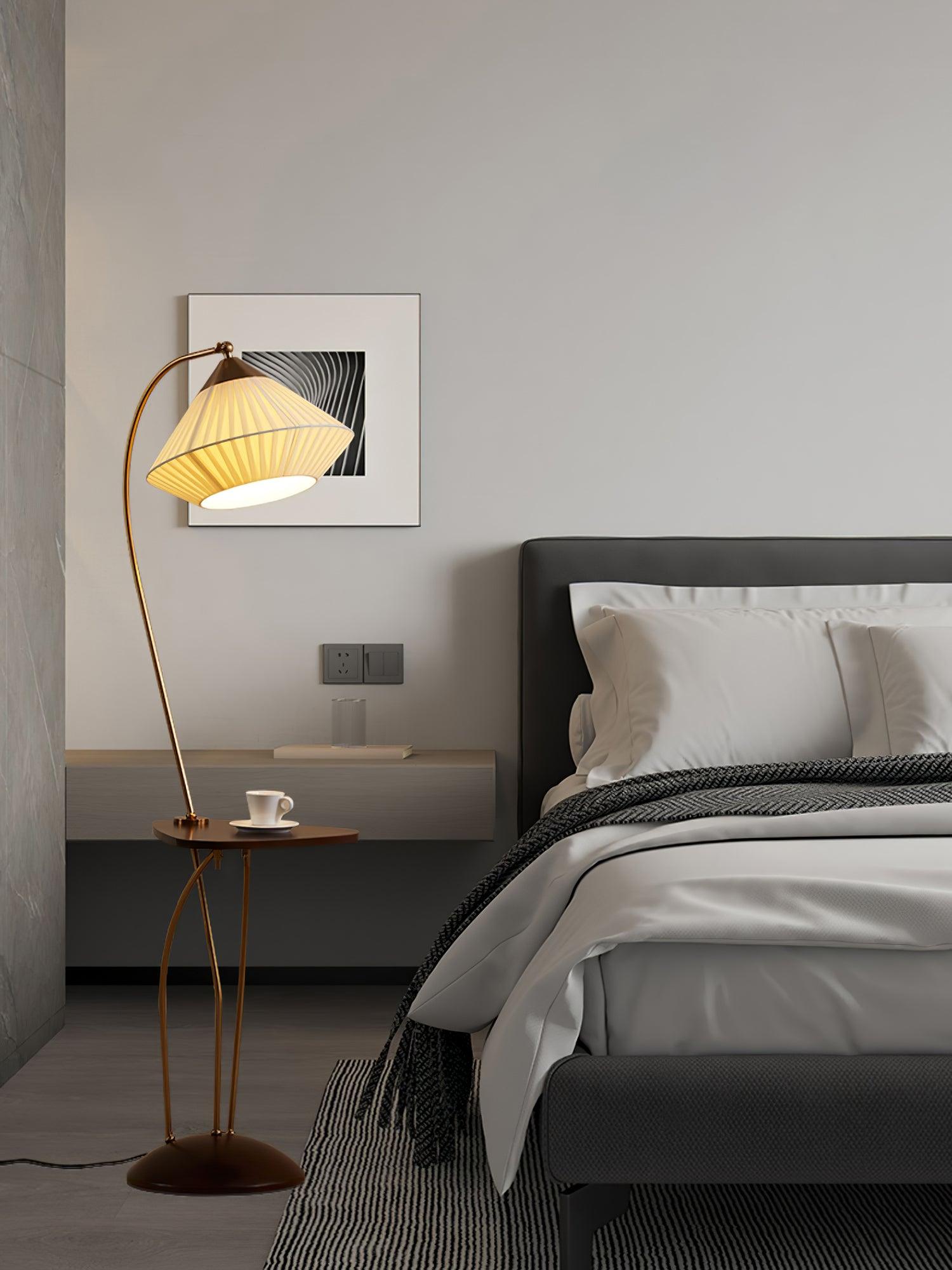 Wide bedroom view of Curve’s glowing pleated shade and cup-topped table beside a bed with white pillows and patterned throw