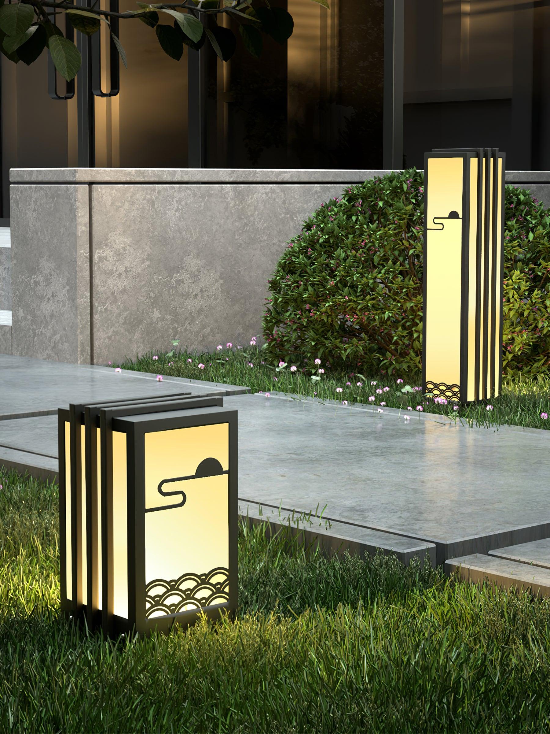 Closer view of short and tall sun-and-wave lanterns on lawn beside stone path, hedge and entrance wall