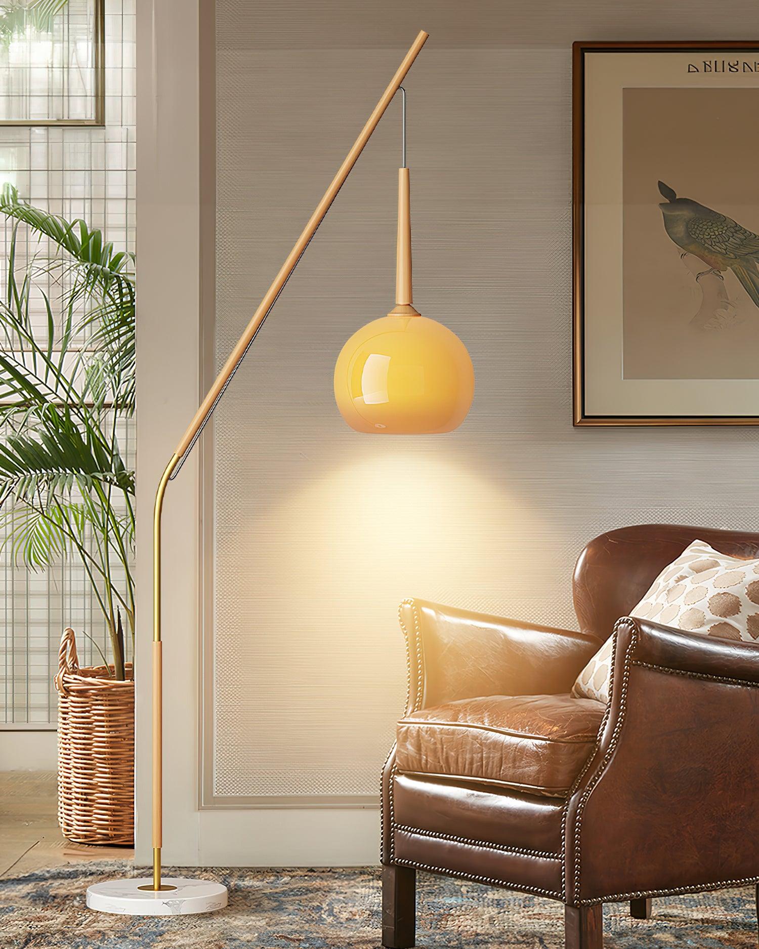 Orange glass shade on an angled natural wood-colour floor lamp beside a brown leather chair, plant basket and bird print