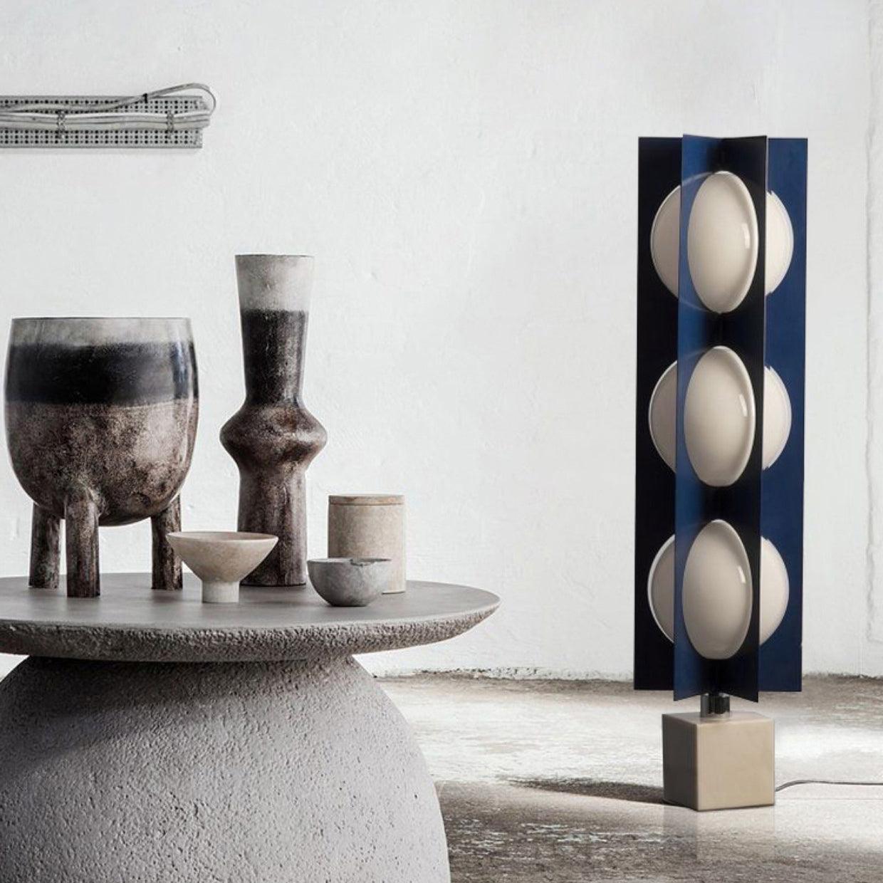 Three-level blue column lamp on the floor beside a round stone table holding vases and bowls