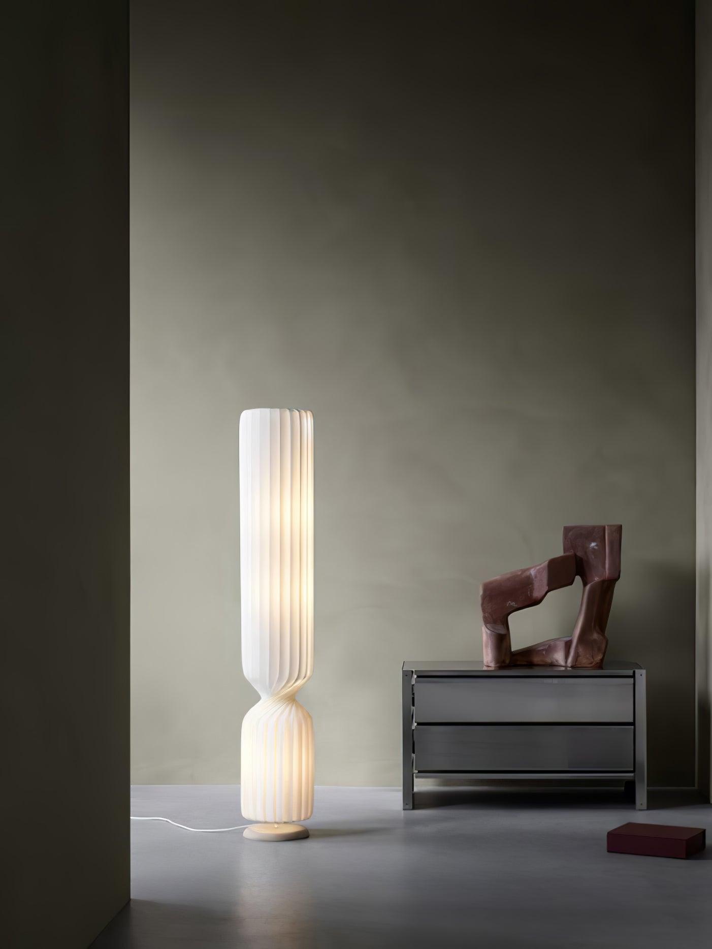 Wide view of lit white pleated lamp beside low gray metal chest and reddish sculpture, red object and cord on floor