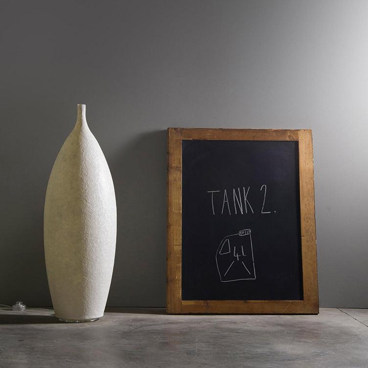 Short white textured bottle light and visible cord beside a wooden TANK 2 chalkboard on grey floor