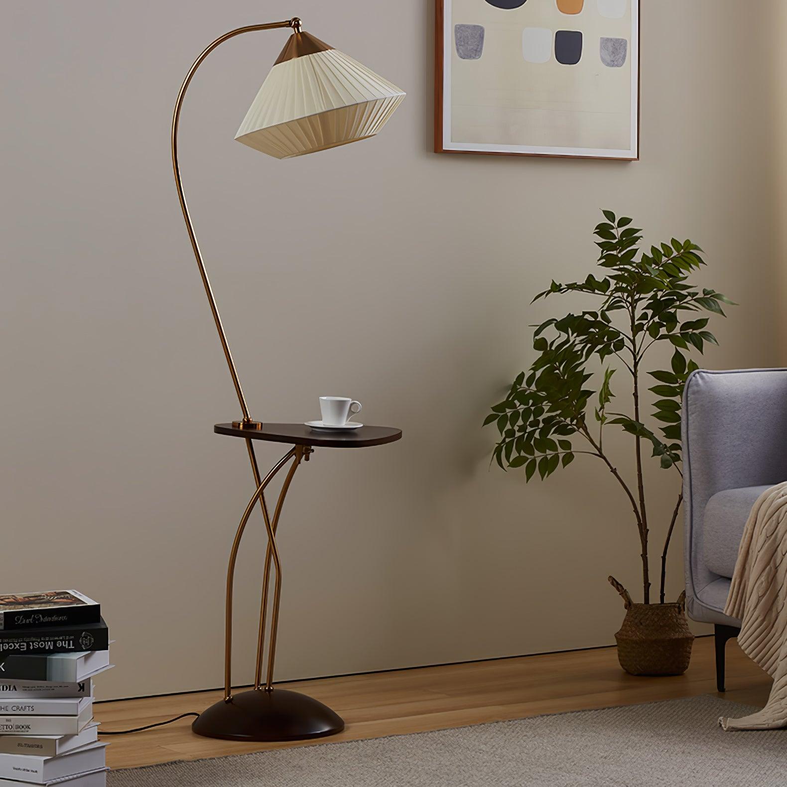 Unlit Curve floor lamp with a beige pleated shade and dark table beside a grey armchair, plant and stacked books