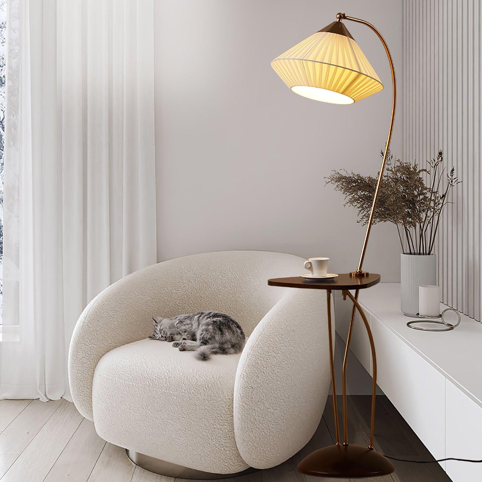 Curve lamp with a lit pleated shade and small table beside a cream chair with a sleeping grey cat near curtains