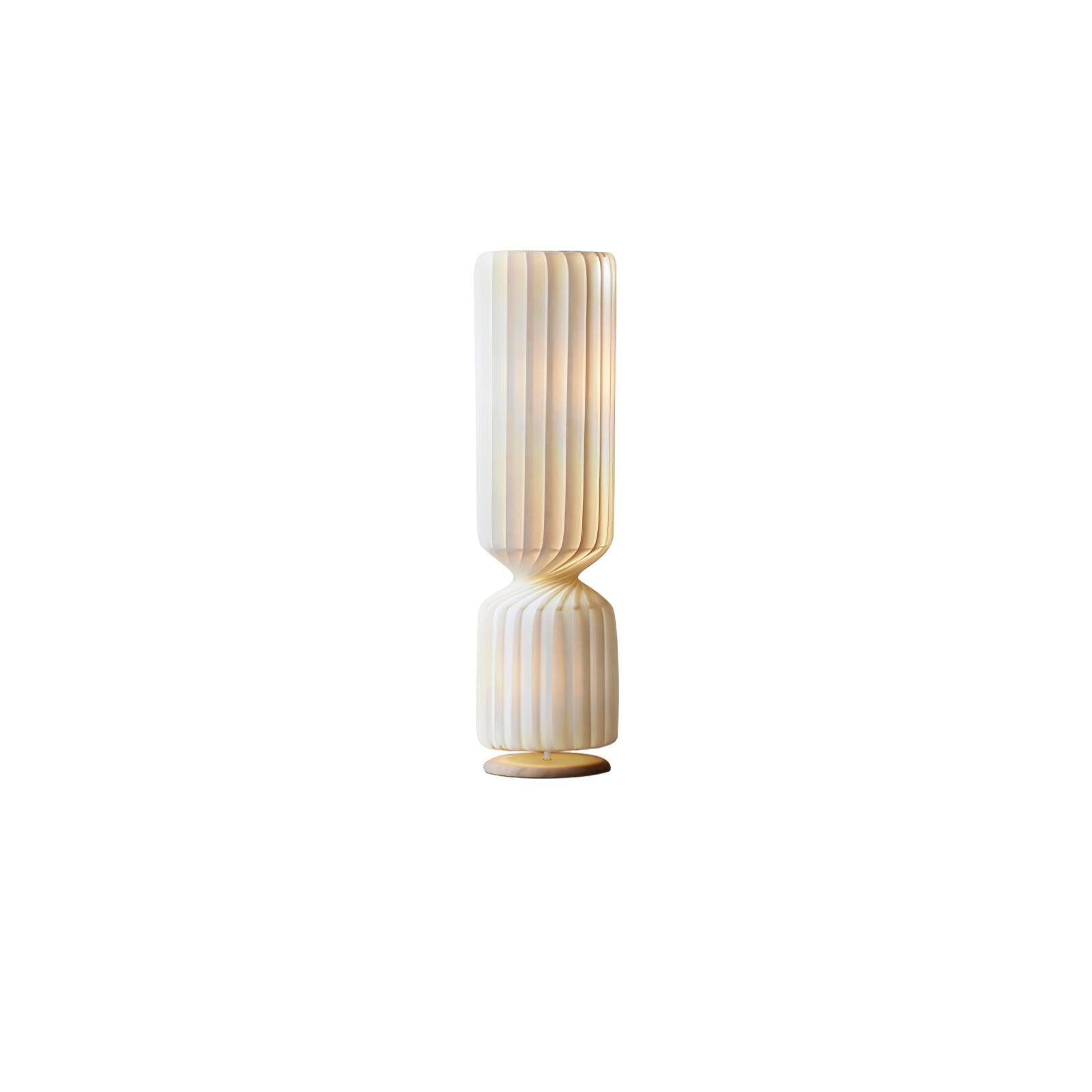 Small isolated lit white twist lamp, pleats curving into a narrow waist over shorter lower section and pale gold-toned base