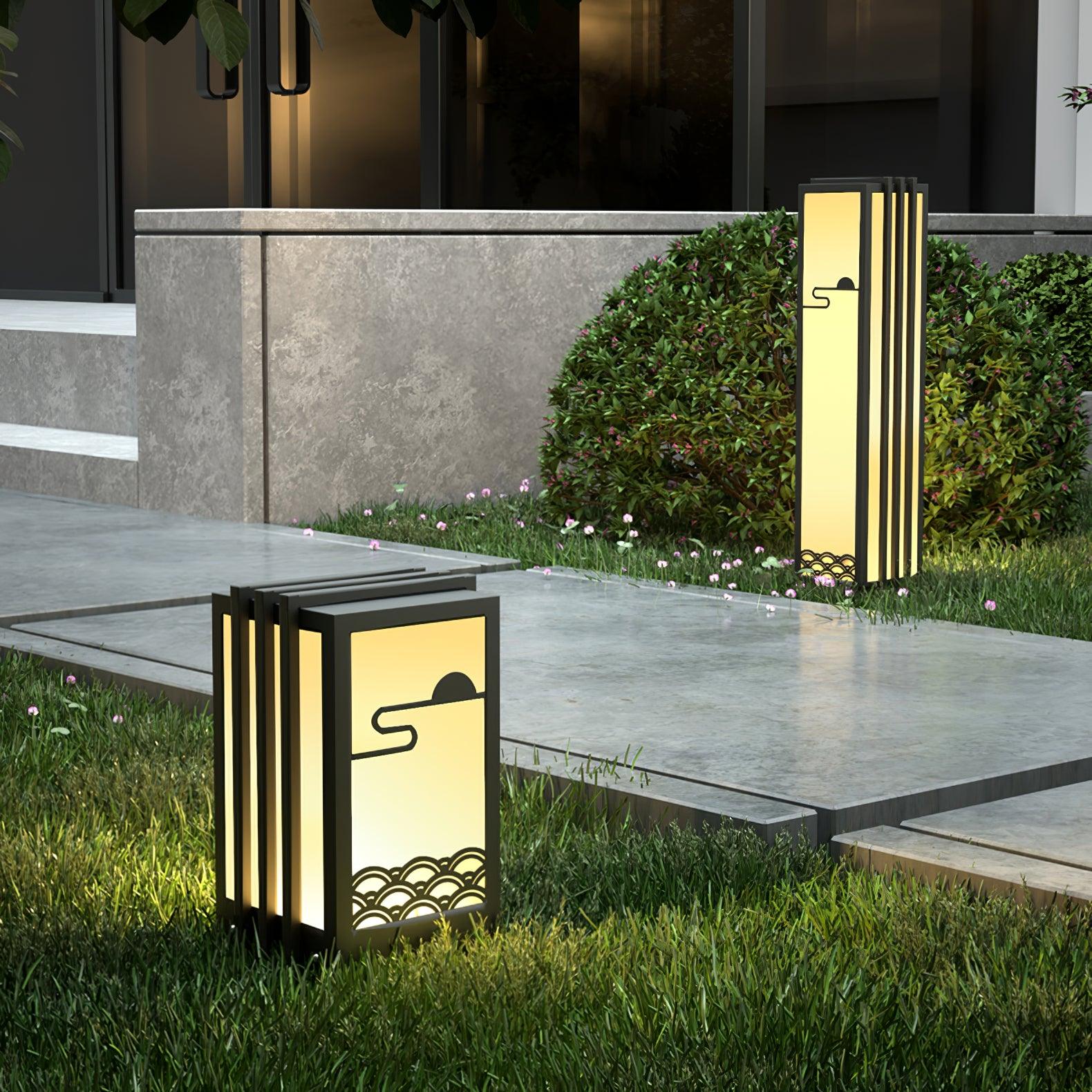 Short broad lantern beside garden paving, with tall slim lantern behind among grass, flowering shrubs and building wall