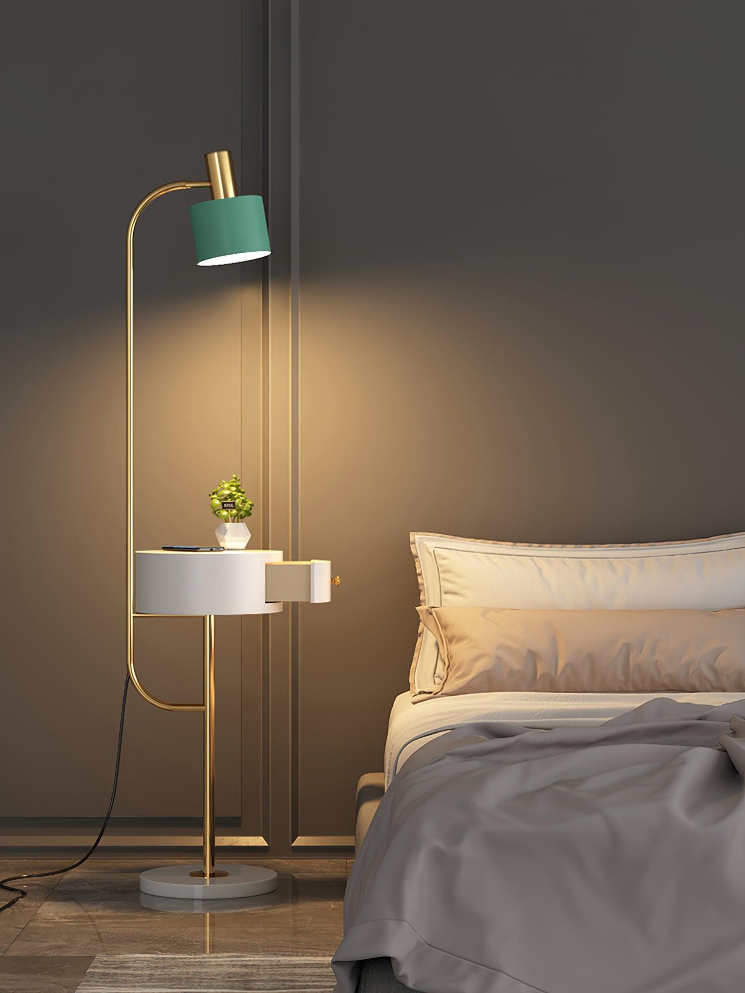 Gold lamp lit beside cream bed and gray wall, green shade above white open drawer table with phone and plant, white base