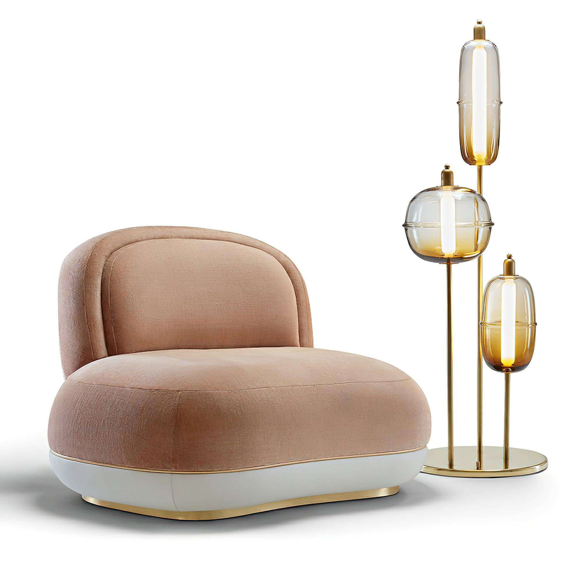 Gold floor lamp with three amber glass shades and circular base beside a curved pink chair on a white background