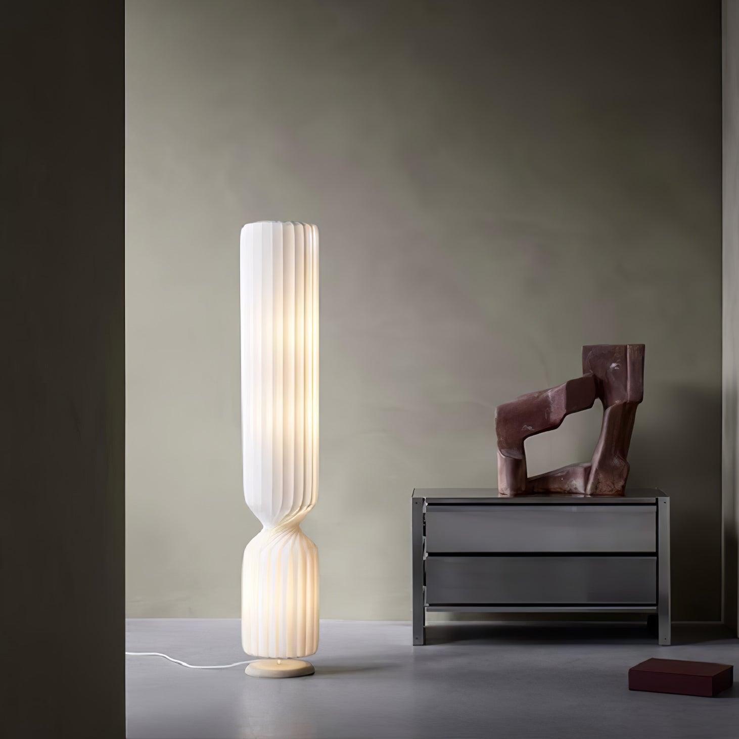 Lit white Twist floor lamp beside gray metal chest and reddish abstract sculpture, with white cord on floor