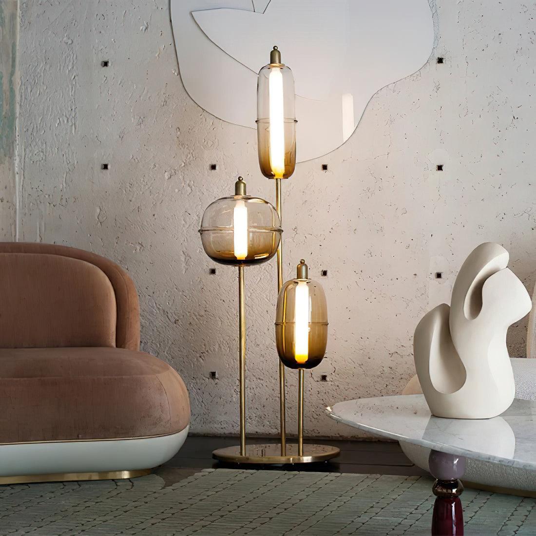 Gold-stem lamp with three amber glass shades beside a pink curved chair, textured wall and white sculpture on a table