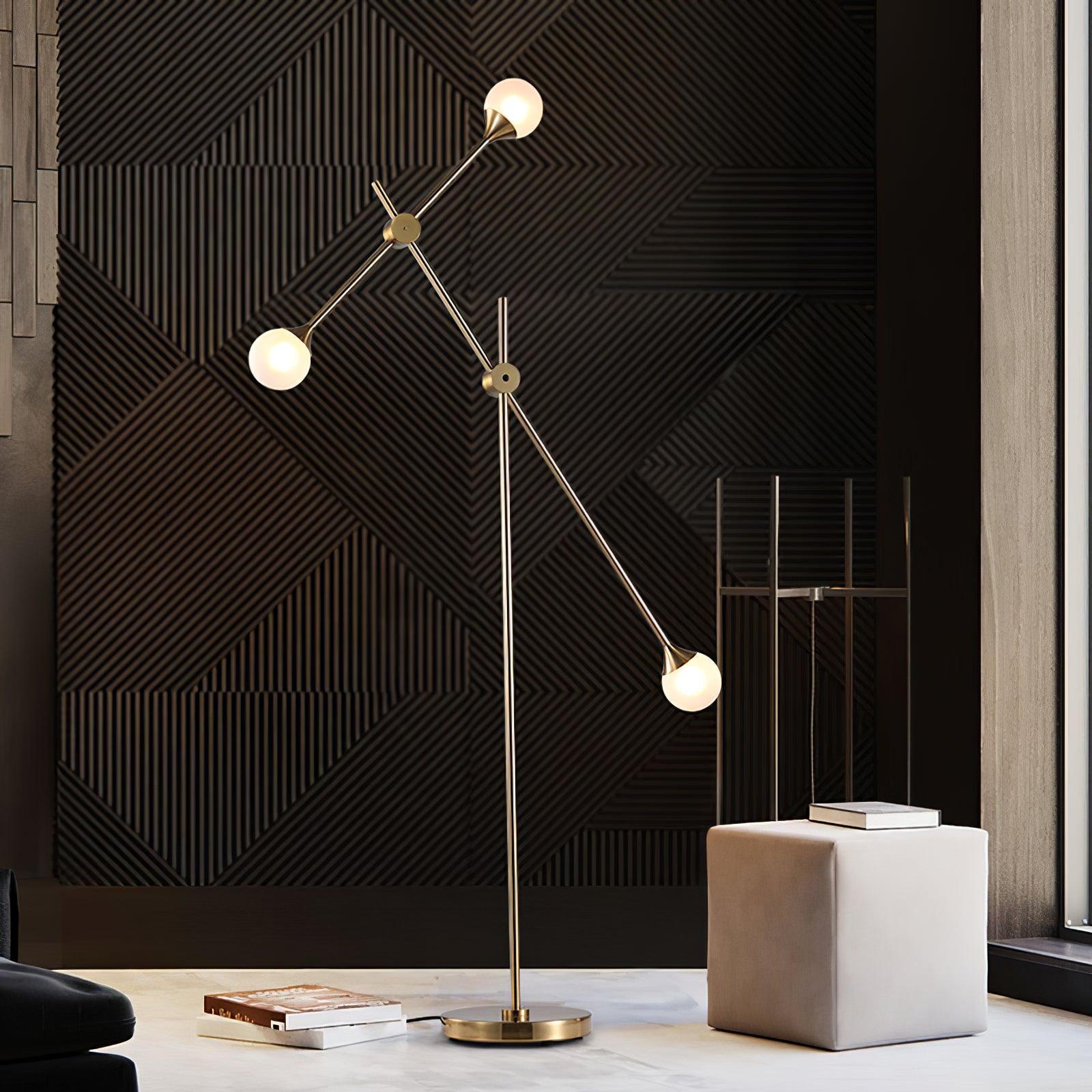 Gold three-globe Kemi lamp against a dark geometric wall, beside a pale cube and books, with a cord by its base