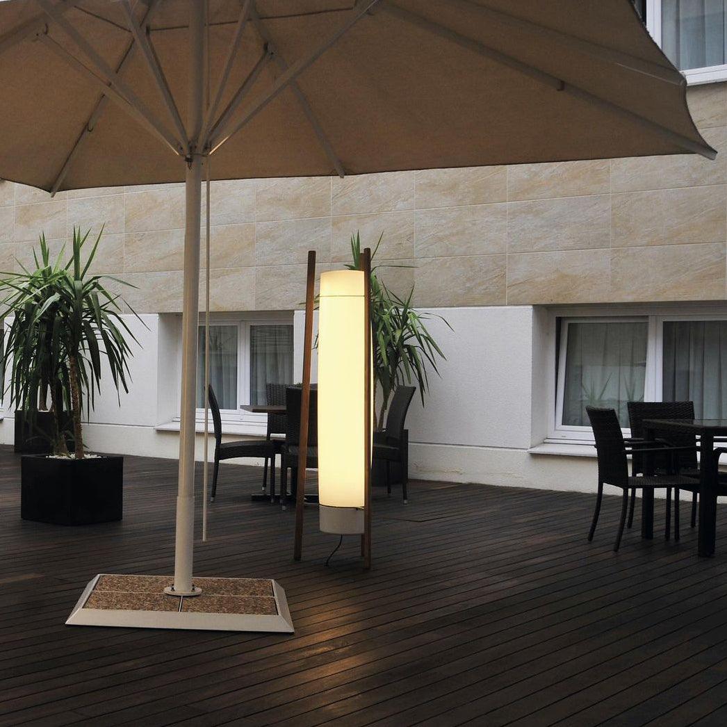 Lit column lamp with wooden supports and grey bottom cap beside a parasol base and dining chairs on a wood deck