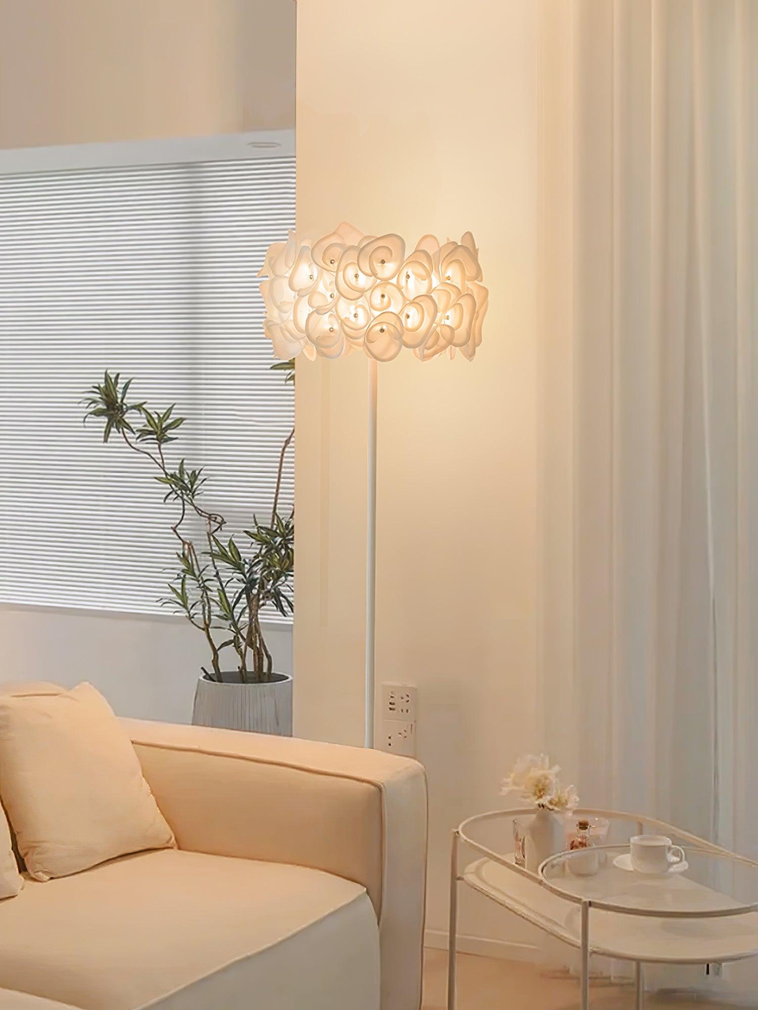 Upright lit white petal lamp by cream sofa, leafy plant, ribbed wall and small round tray table with cup