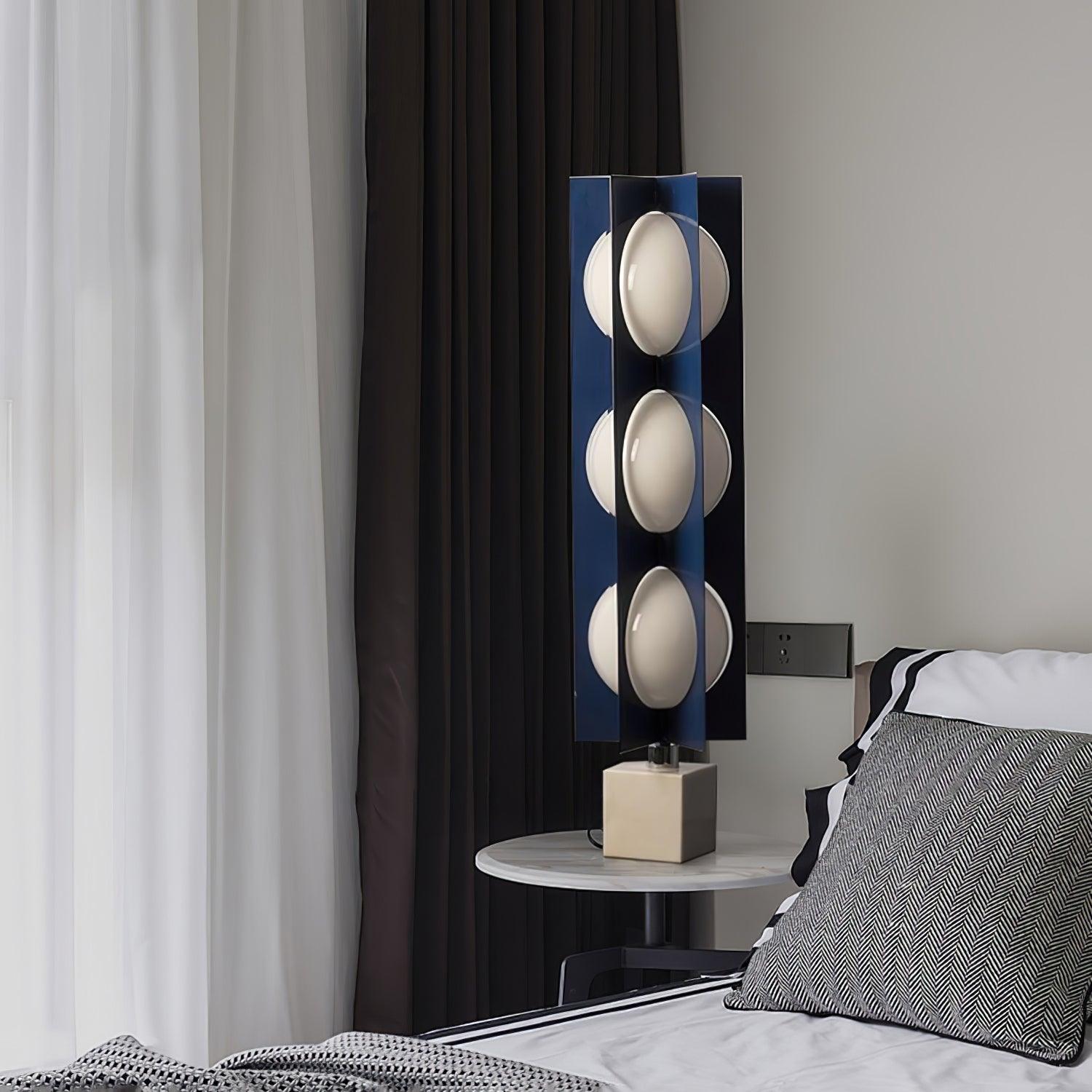 Wider view of the three-level blue column lamp on a bedside table near white and dark curtains